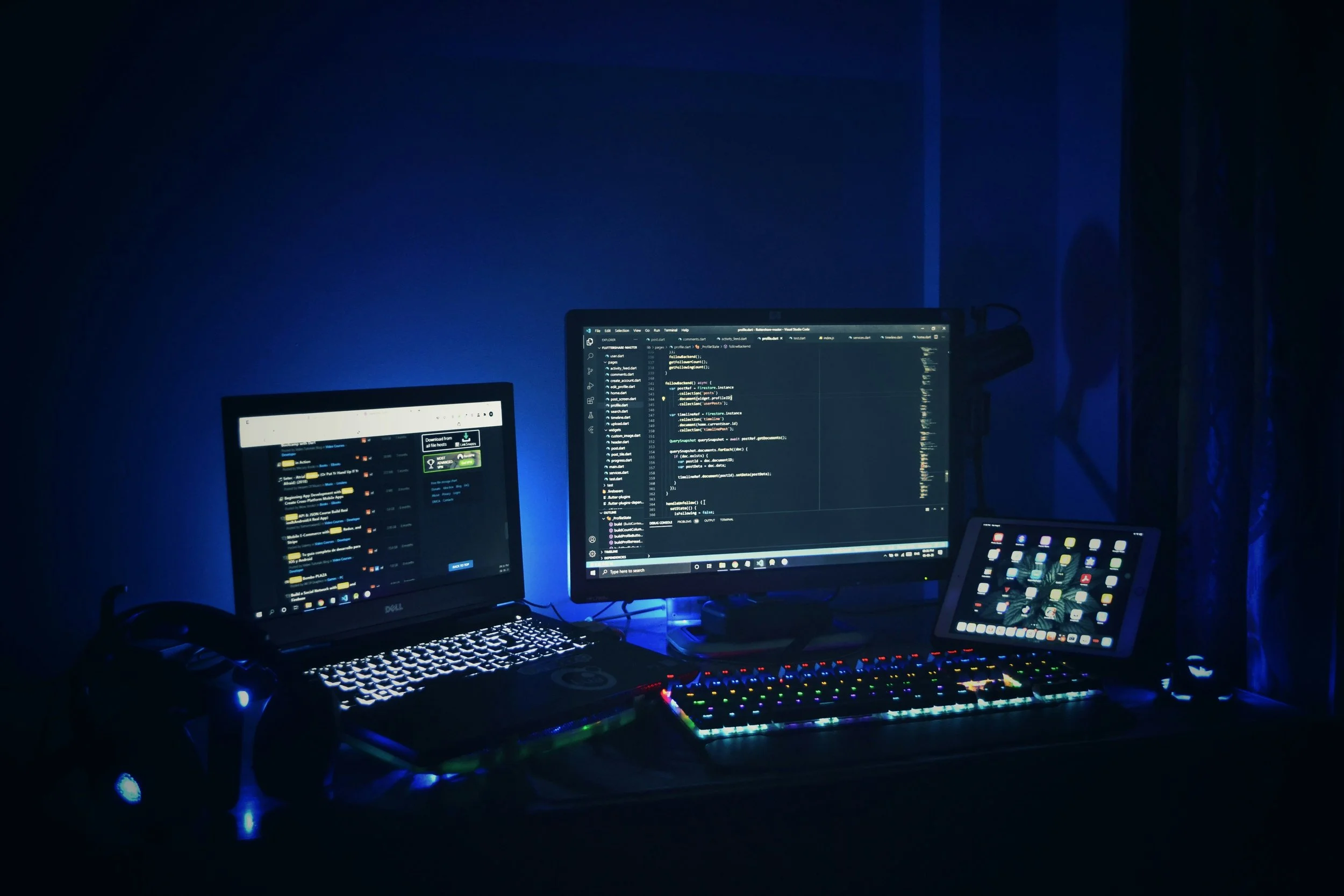 A computer setup with multiple screens, including a laptop, monitor, and tablet, displaying coding and digital interfaces in a dark room with blue ambient lighting.
