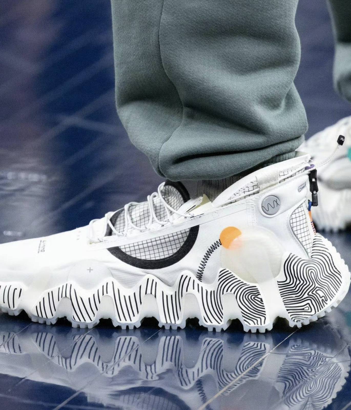 Close-up of a futuristic white sneaker with black and orange accents, featuring a textured sole with black wavy lines, worn with green pants on a shiny blue surface.