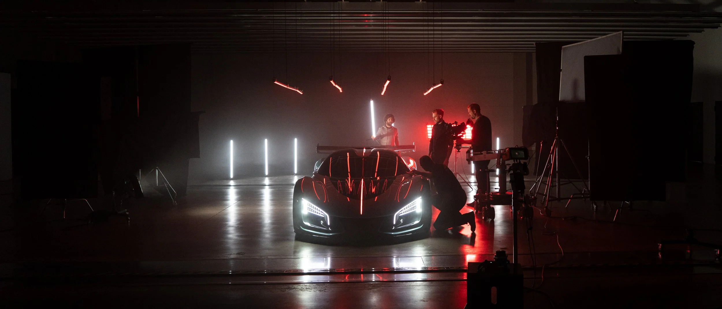 A black sports car on a studio set with three film team members working around it, illuminated by red and white lights, with equipment and lighting setups visible in a dark, industrial-style space.