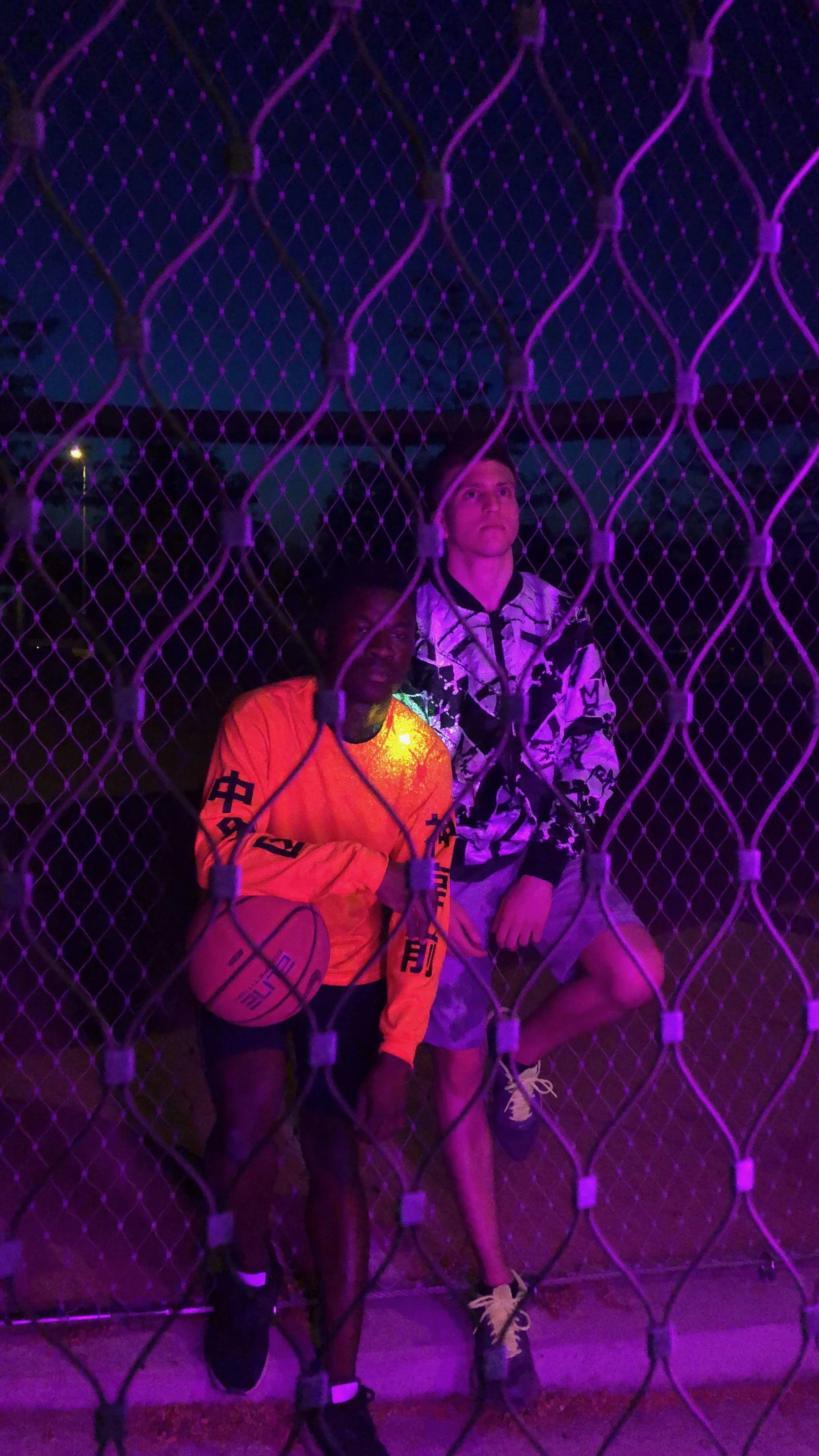 Two men behind a chain-link fence at night with purple and pink lighting. One man is holding a basketball and wearing an orange shirt, the other man is wearing a patterned black and white jacket and shorts.