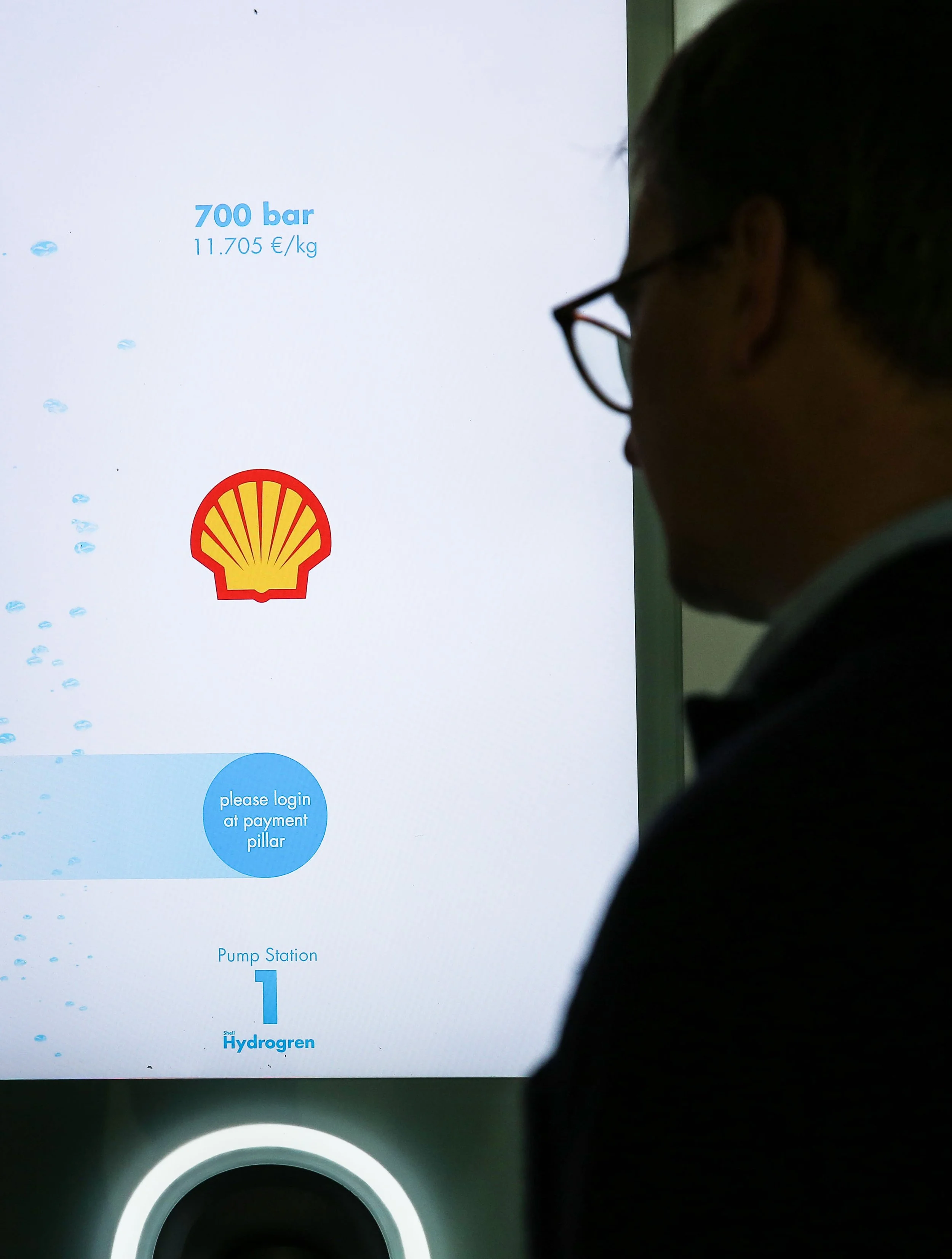 Person looking at a Shell hydrogen fuel pump payment screen displaying 700 bar and 11.705 €/kg, with a Shell logo, instructions to login at payment pillar, and a pump station labeled 'Hydrogen 1'.