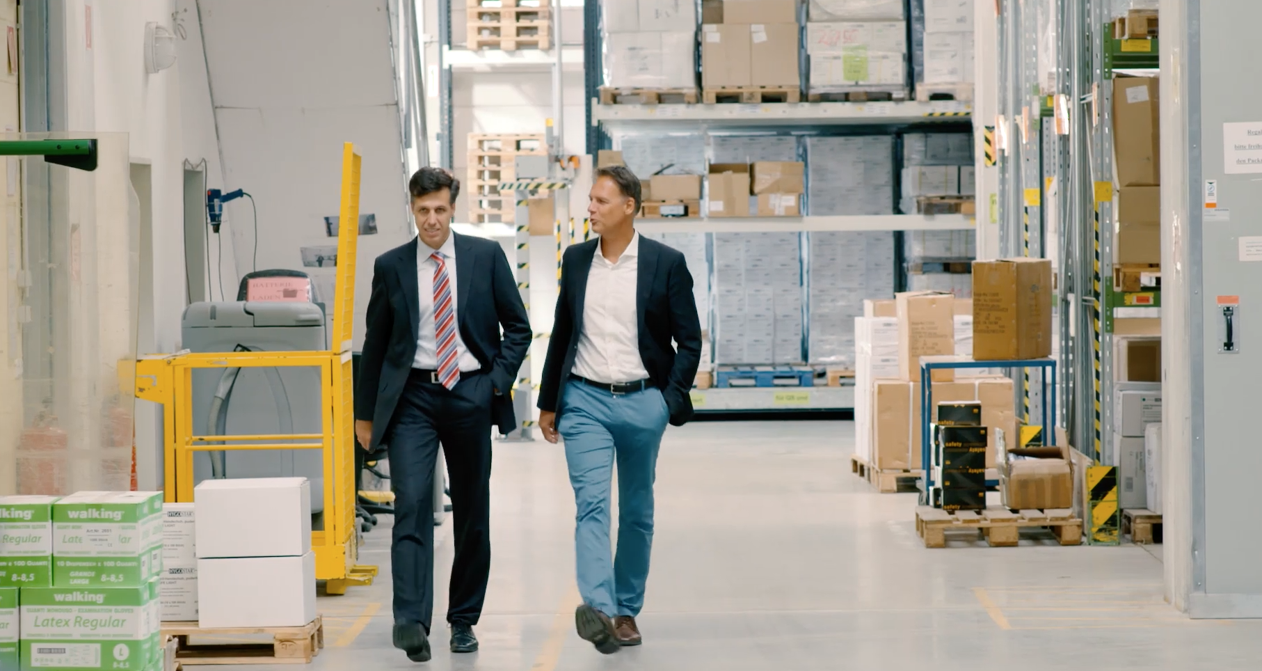 Two CEOs in suits walking and talking inside a warehouse with shelves of boxes and pallets.