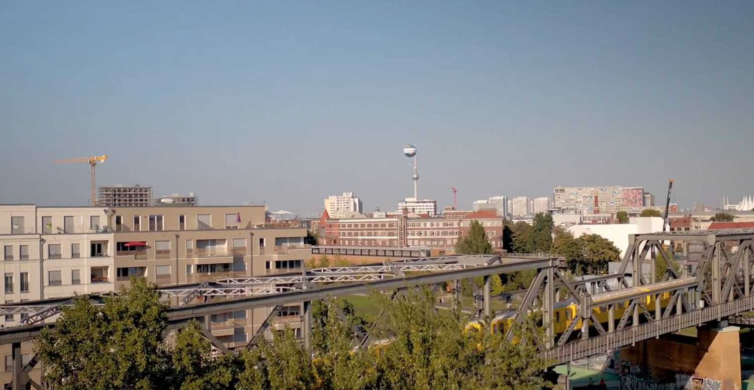 Berlin cityscape view with buildings, trees, a cable car train on a track, and a television tower in the background on a clear day.