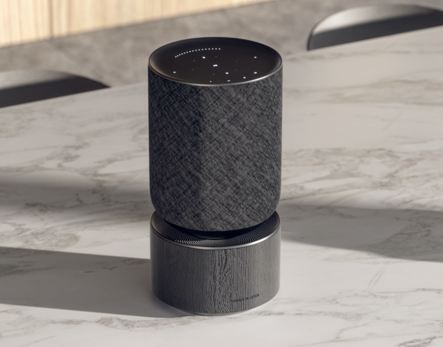 A smart speaker by B&O with a fabric exterior on a marble surface, with a dark top and a cylindrical base.