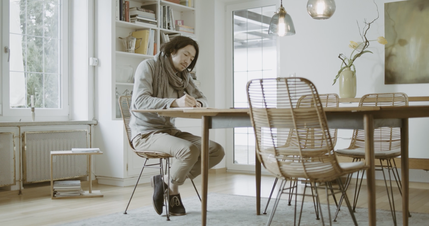 An Asian man sitting at a wooden dining table writing on paper in a bright, modern kitchen with large windows, bookshelves, and a decorative plant in a vase.