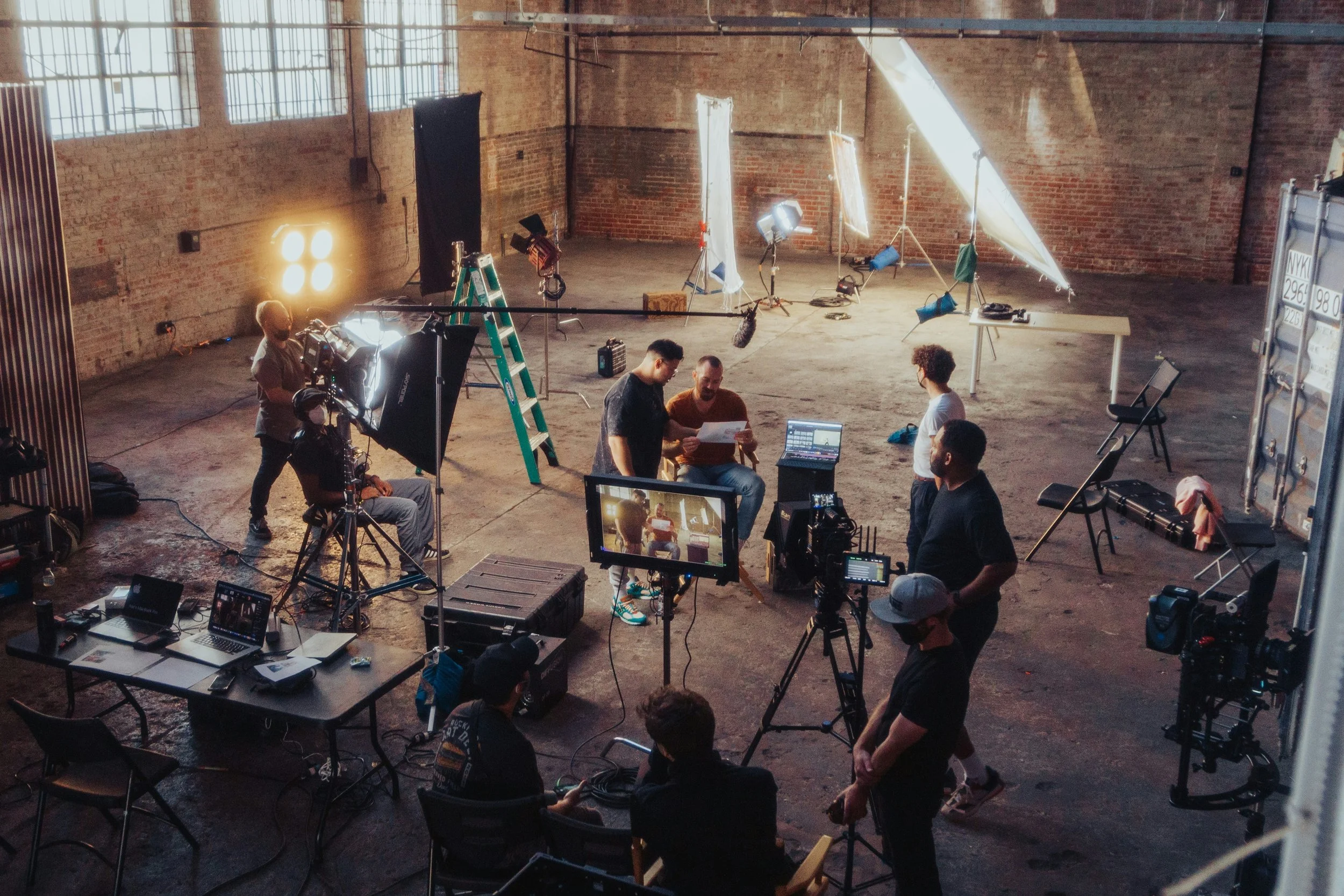 Film crew setting up a shoot in a spacious industrial-style studio with brick walls, large windows, and studio lighting.