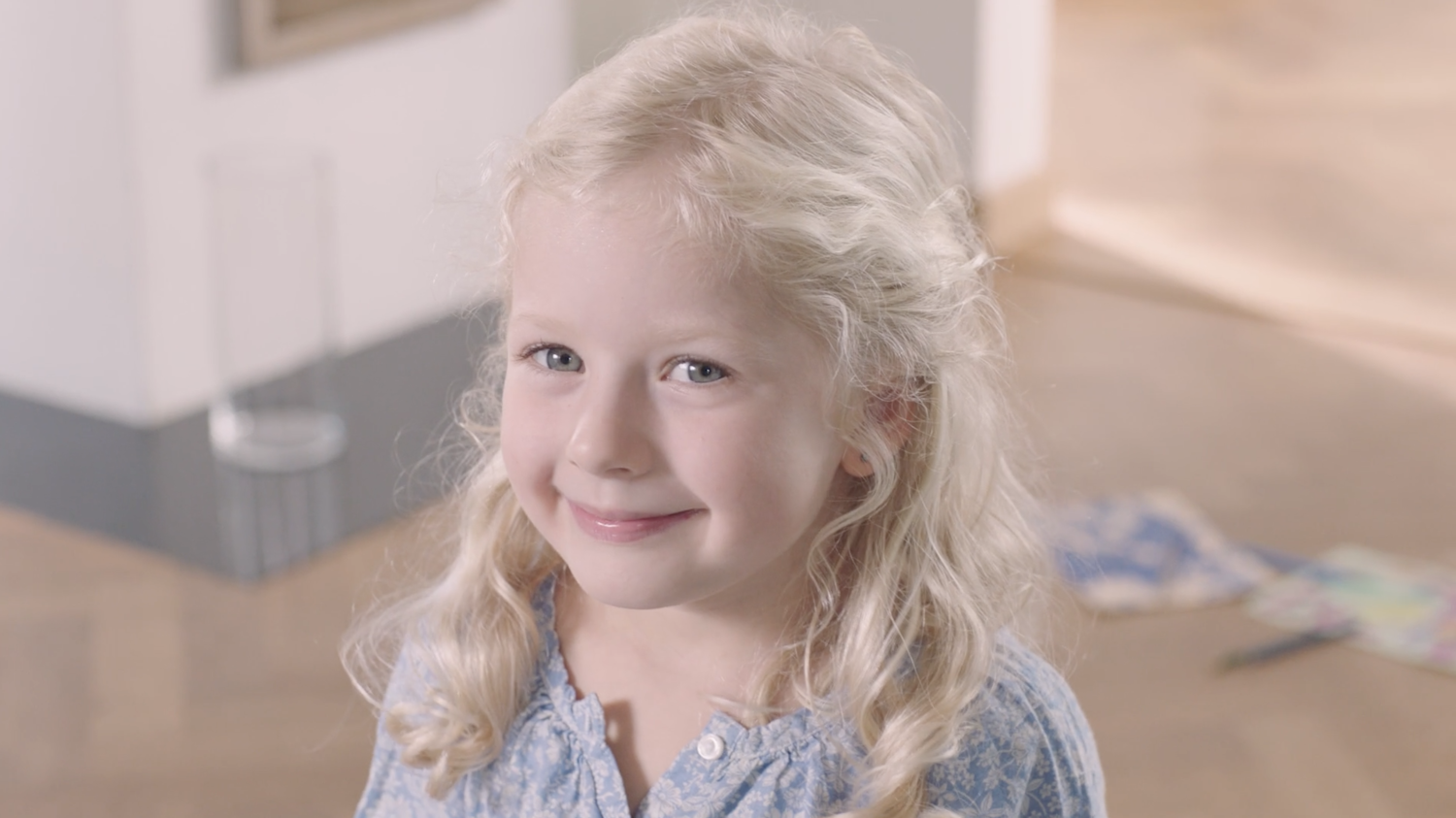 A young girl with blonde curly hair and blue eyes smiling at the camera indoors.