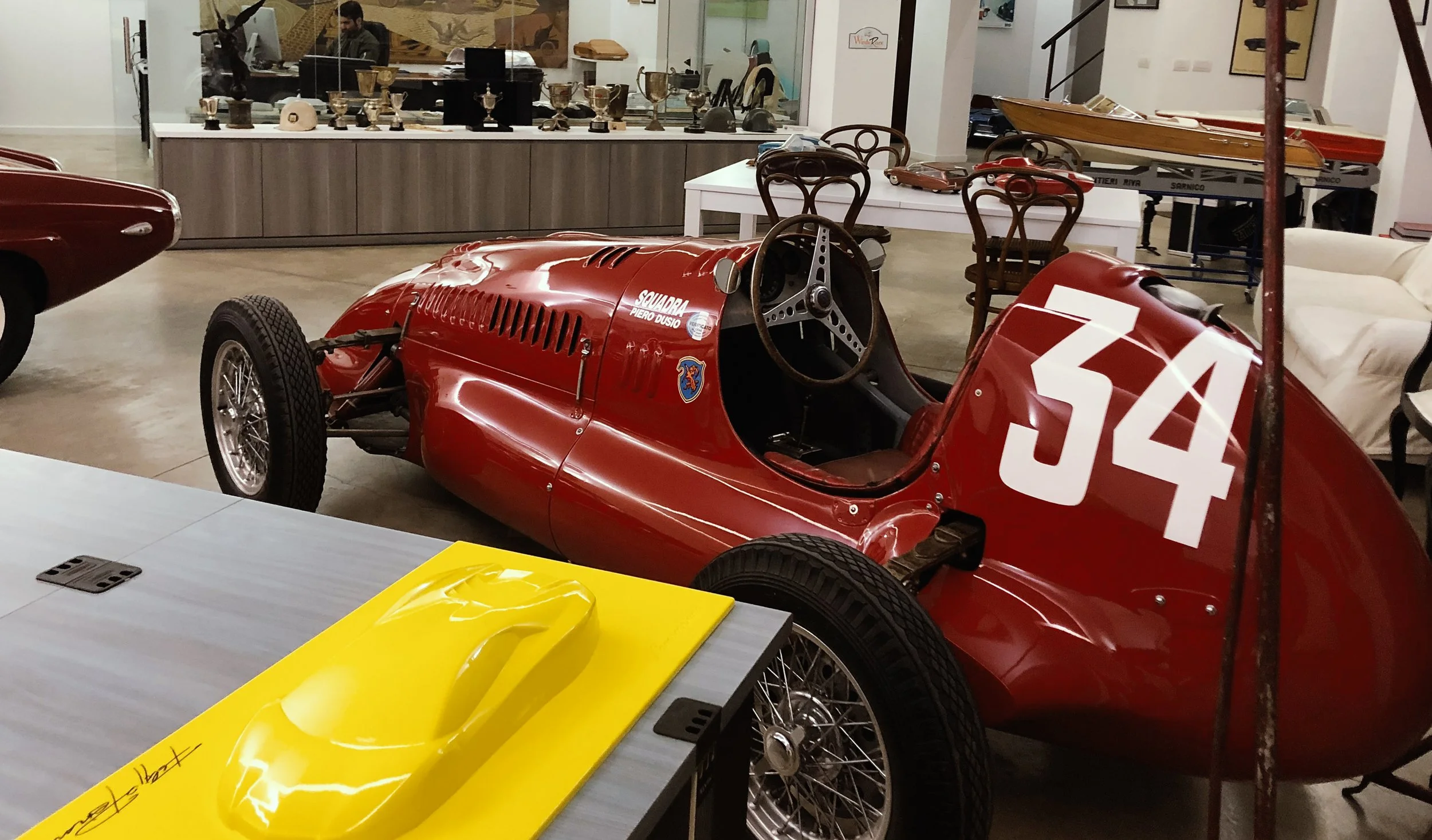 A vintage red race car with the number 34 on the side, on display in an indoor showroom surrounded by trophies and memorabilia.