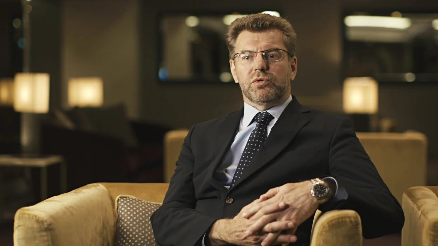 A middle-aged manager with glasses, a beard, and a mustache wearing a dark suit and tie sitting on a yellow sofa in a warmly lit room, looking to the side with a serious expression in an interview.