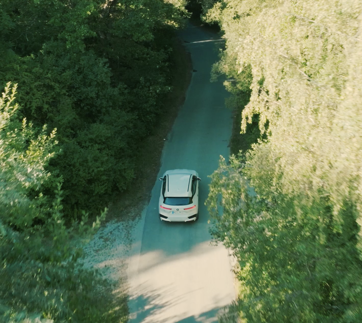 Overhead view of a white BMW iX driving on a narrow, winding forest road surrounded by green trees.