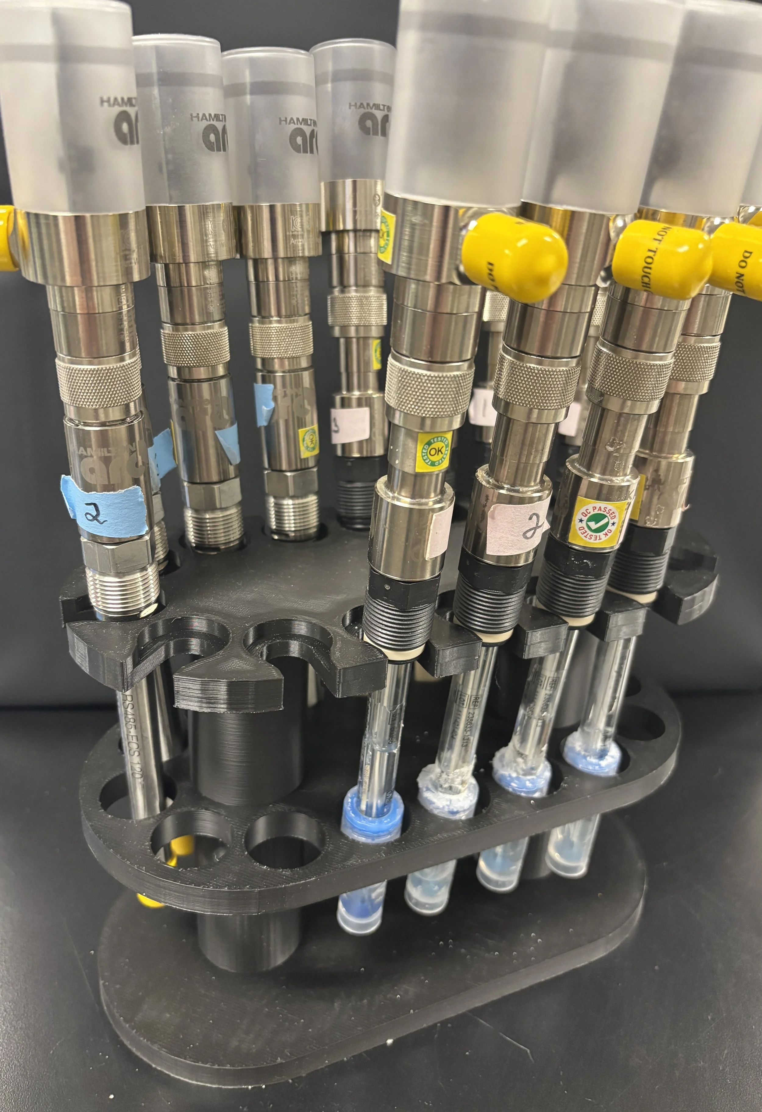 A rack holding multiple medical or laboratory syringes with attached needles, some labeled with color-coded tags and stickers, on a black surface.