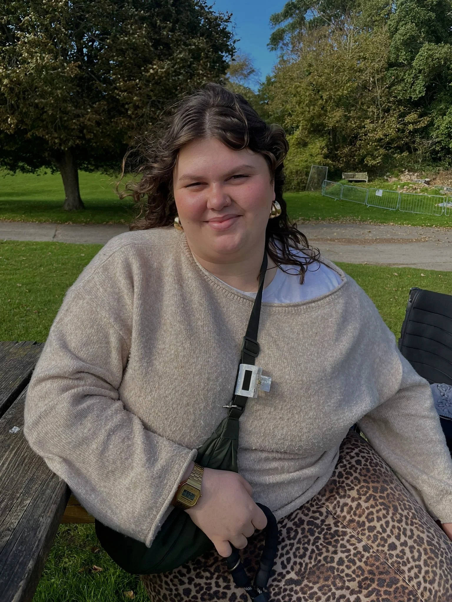 A woman sitting on a wooden bench outdoors in a park, wearing a beige sweater, a leopard print skirt, a gold watch, and earrings, with trees and greenery in the background.