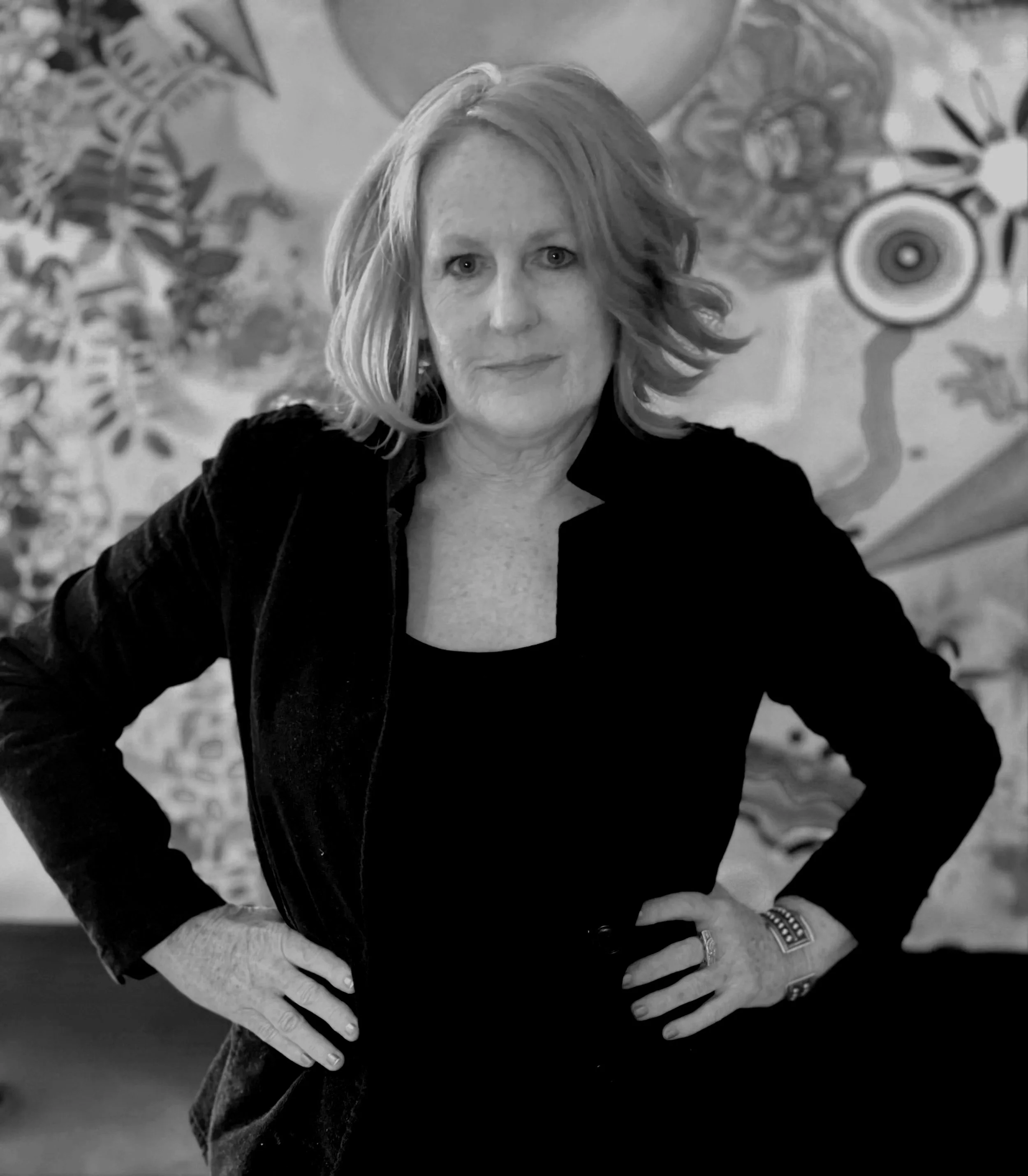 A woman with shoulder-length wavy blonde hair, wearing a dark blazer and black top, stands with hands on hips in front of a colorful abstract mural.