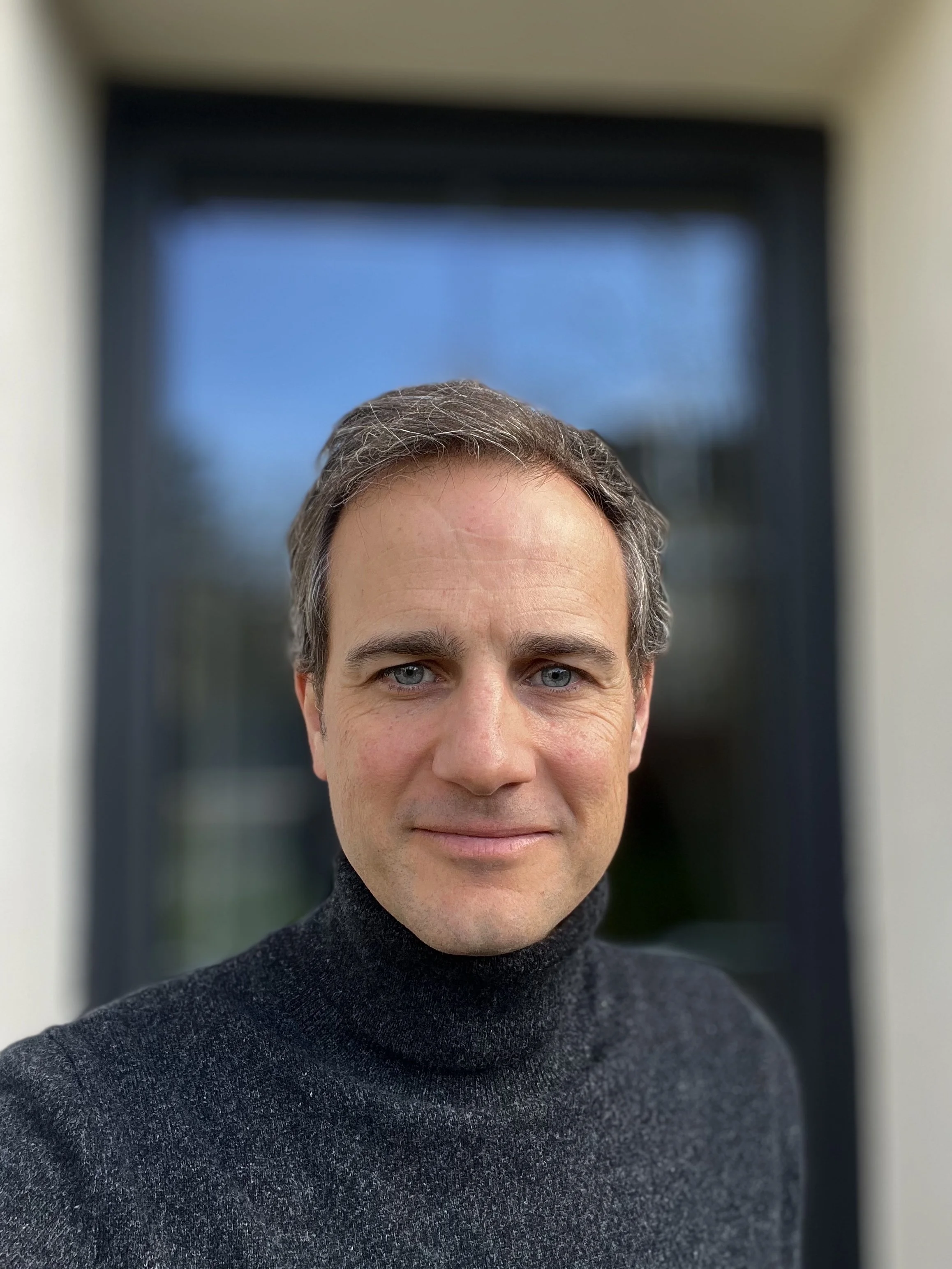 Portrait d'un homme avec des cheveux gris, portant un pull noir, devant une porte en verre.