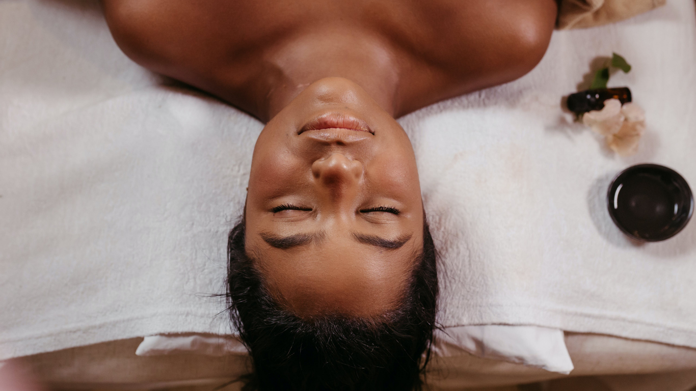 A woman with closed eyes lying on a massage table during a facial treatment with skincare products nearby.