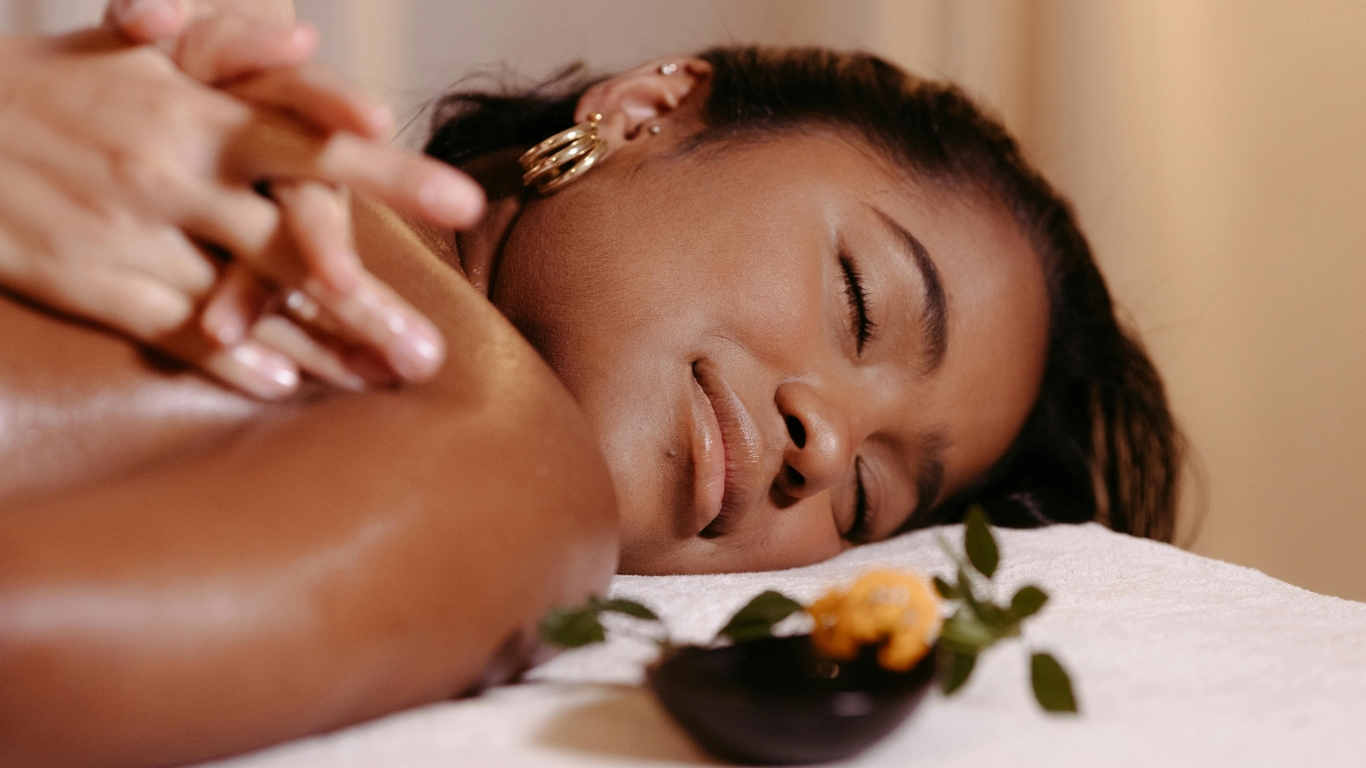 A woman lying face down on a massage table, receiving a massage, relaxing with eyes closed, with a small dish of herbs and flowers nearby.