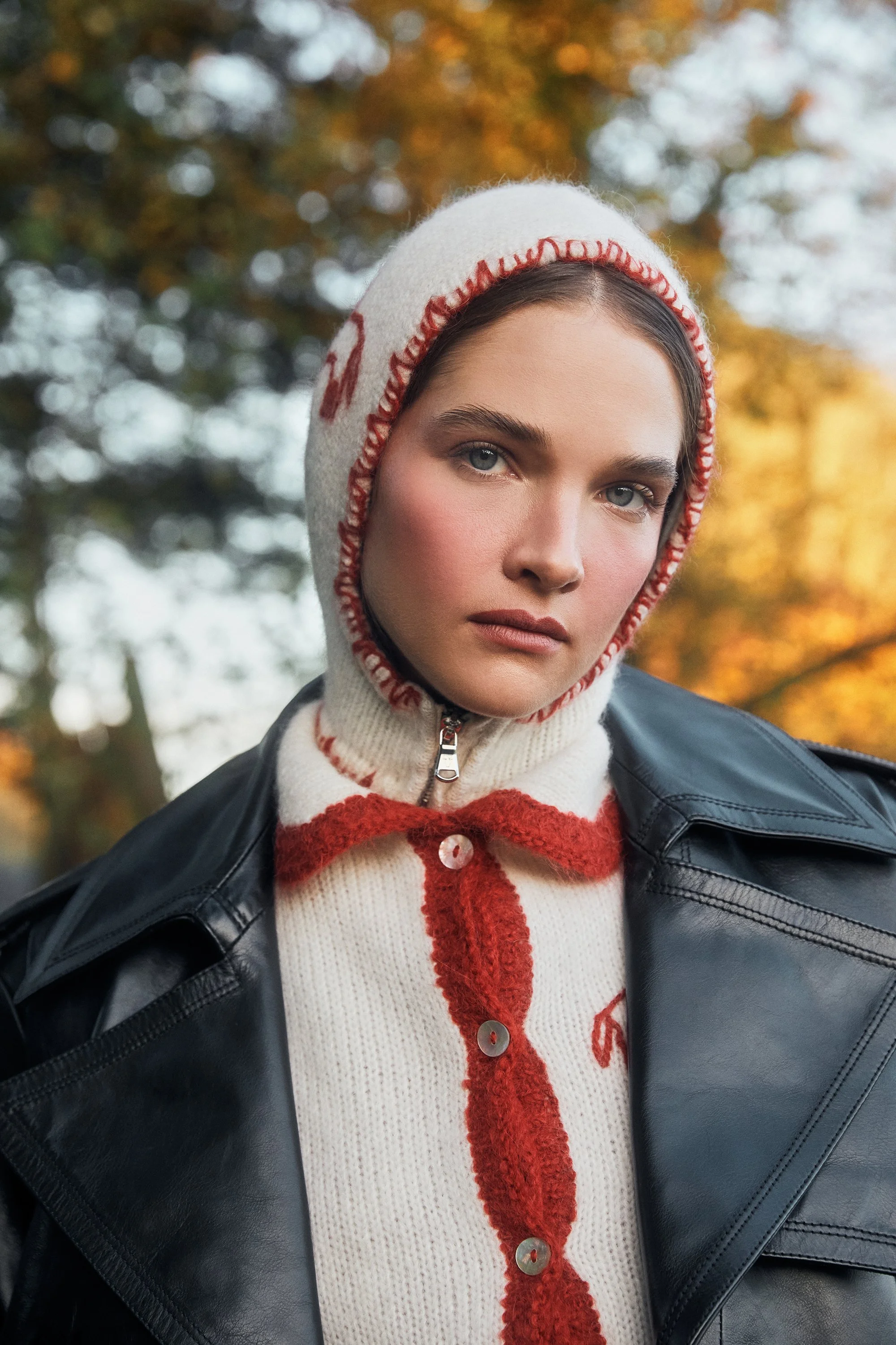 A woman wearing a knit hoodie with red and white details and a black leather jacket, outdoors with trees in the background during autumn.