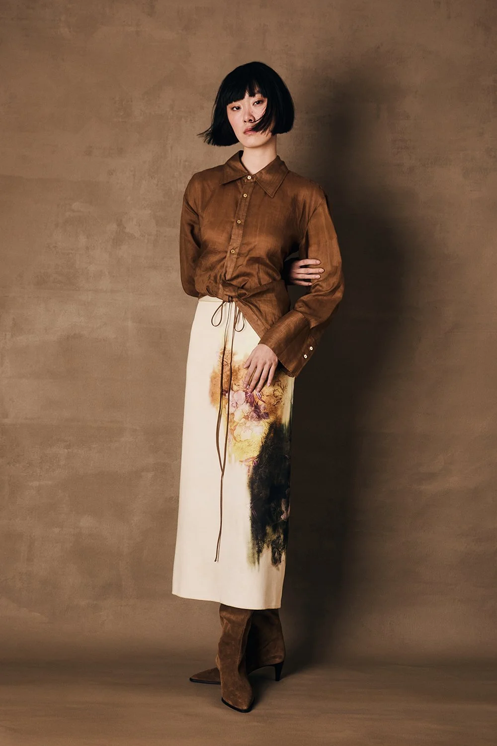 A woman with short black hair wearing a brown button-up shirt tied at the waist, a long cream-colored skirt with an artistic floral and abstract design, and brown boots standing against a plain brown background.