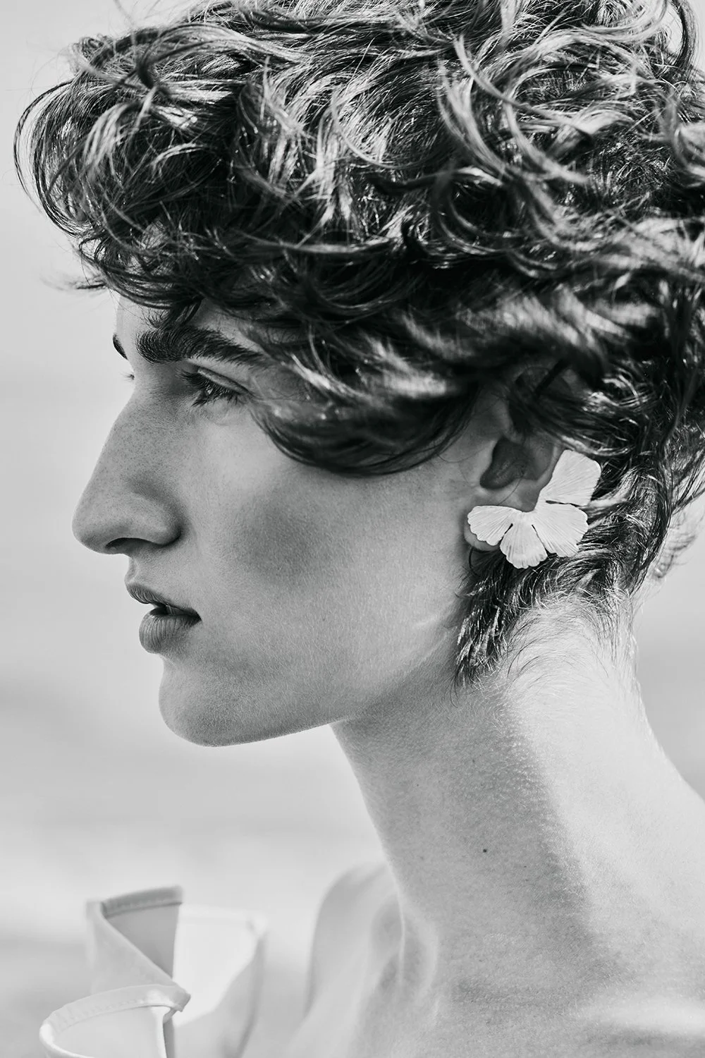A black and white close-up profile of a young person with curly hair wearing a flower earring.