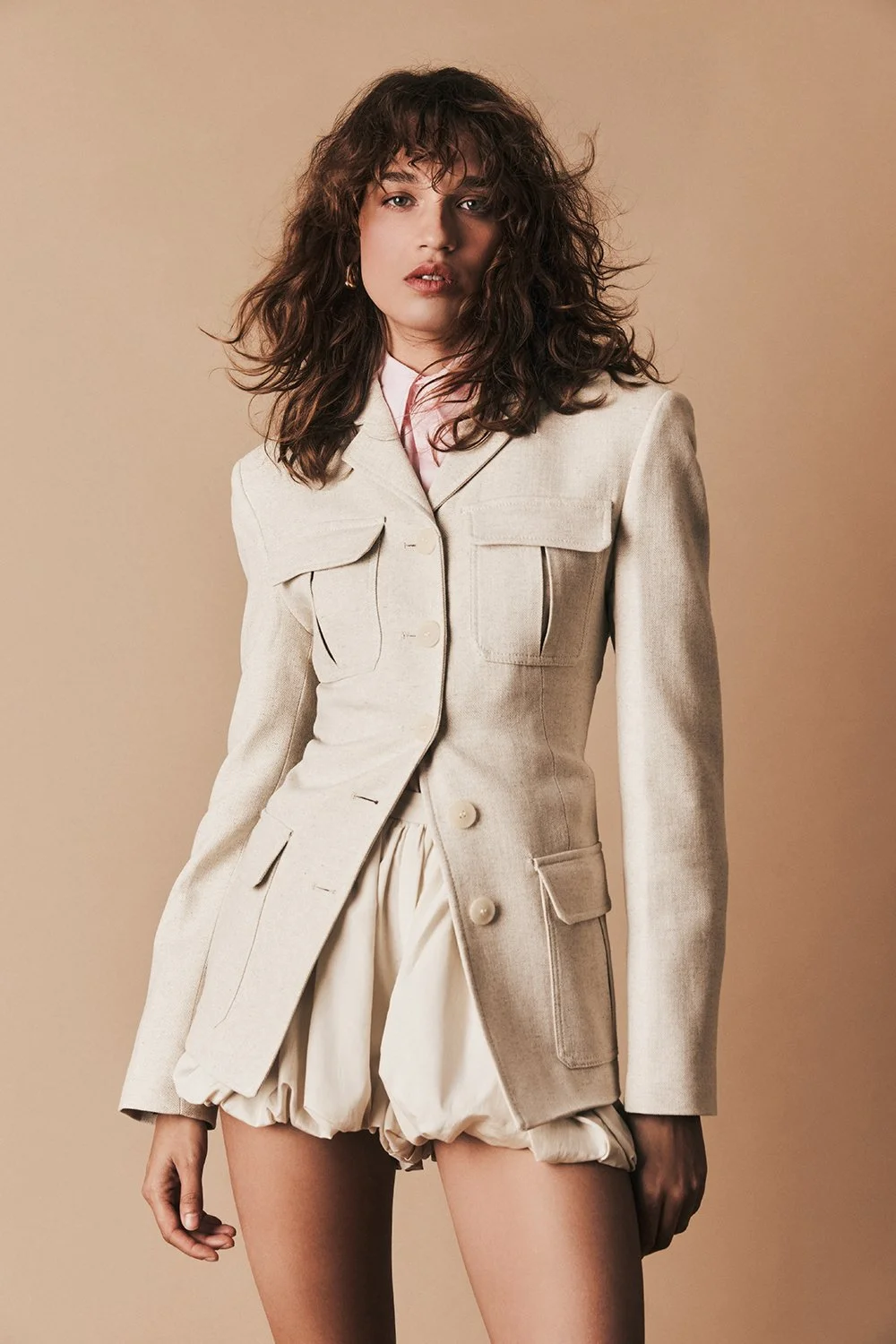 A woman with wavy brown hair wearing a beige jacket over a pink shirt and beige puffed shorts, standing against a neutral background.