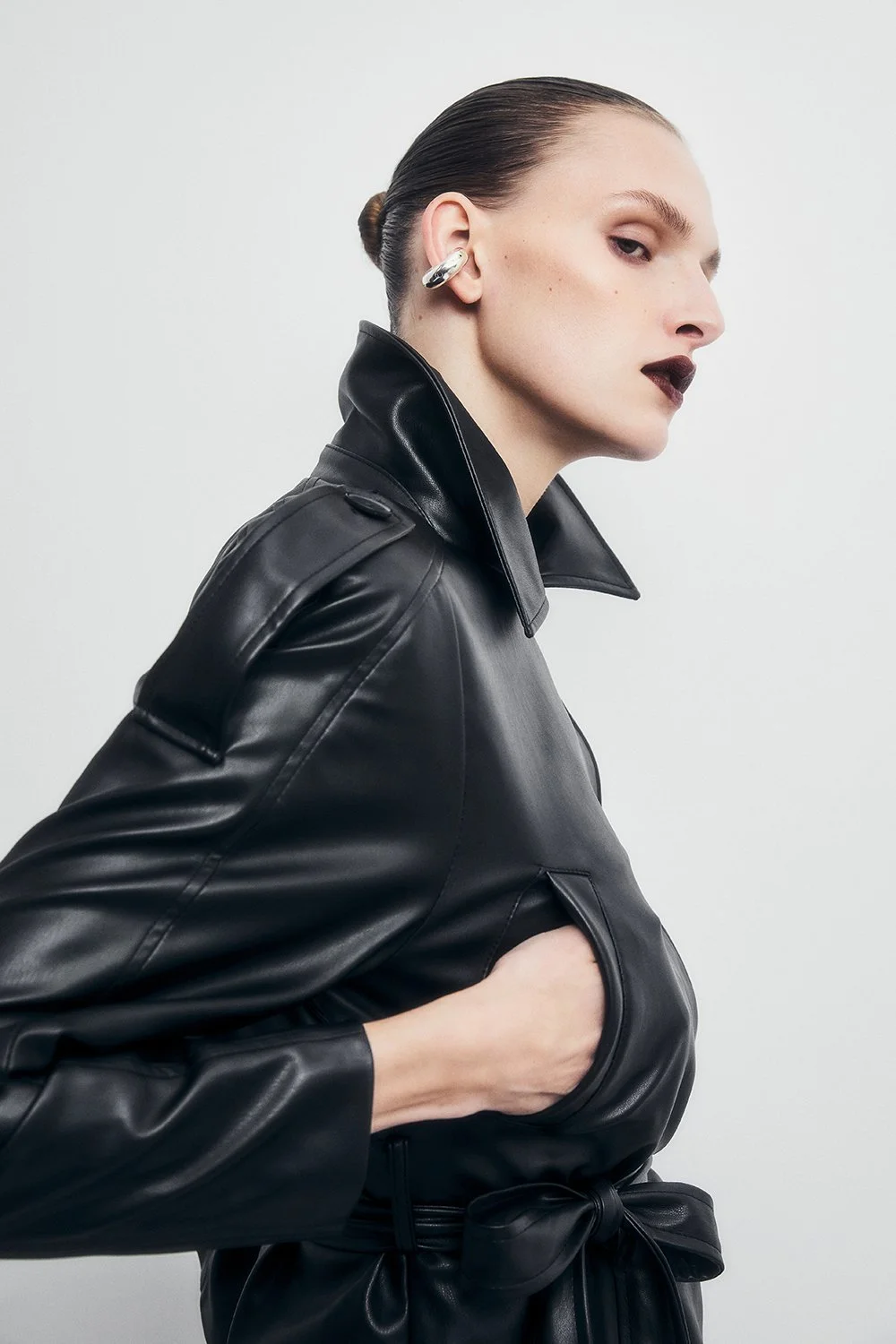 A woman with slicked-back hair, wearing dark lipstick, silver earrings, and a black leather jacket with a belt, standing against a plain white background.