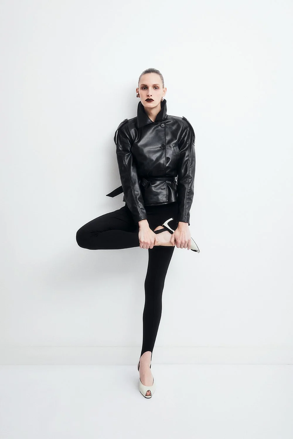 A woman dressed in black standout clothing, standing against a white background, posed with one leg raised and holding her high heel shoe.