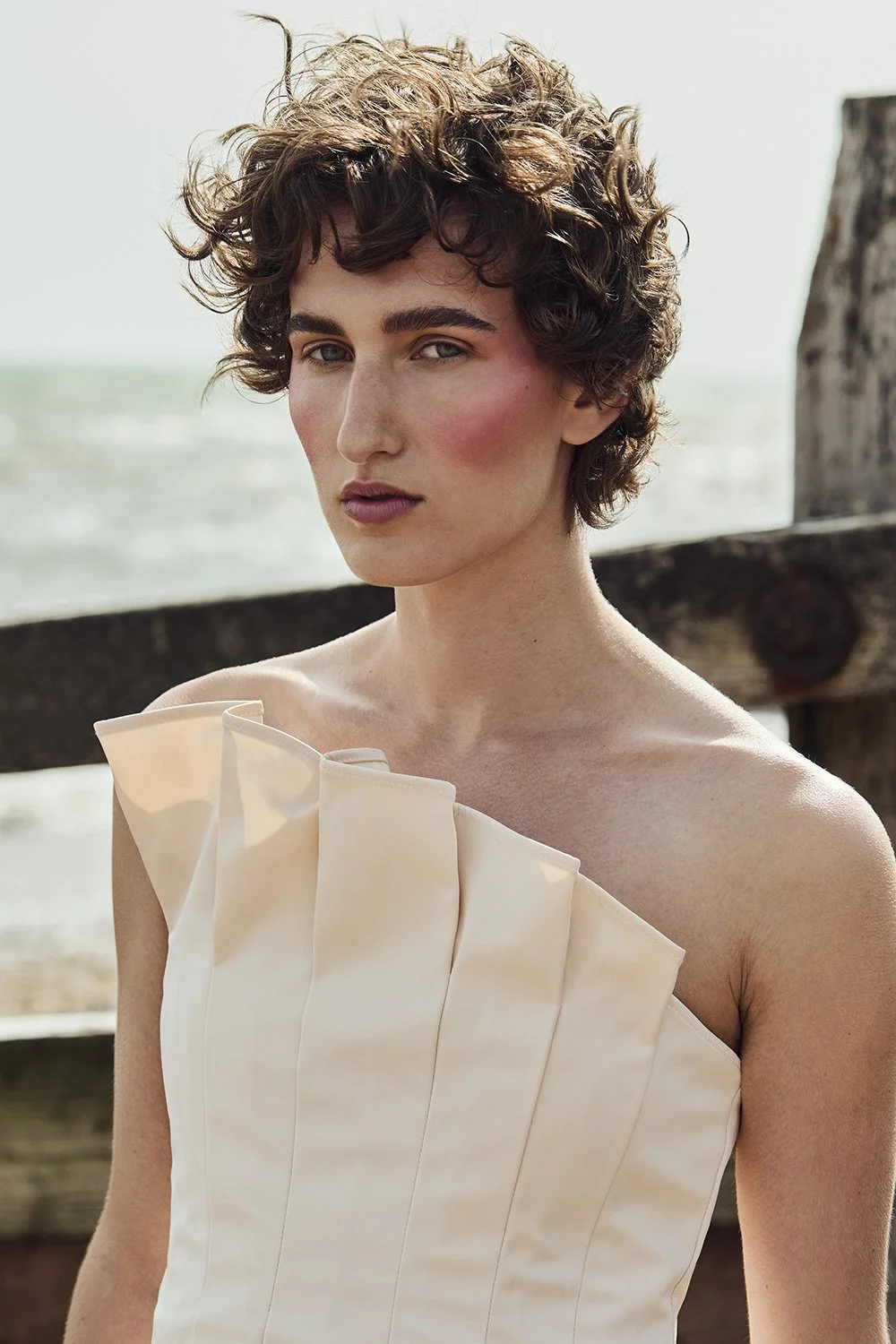 A woman with short curly brown hair and fair skin poses outdoors near a wooden railing, wearing a cream-colored strapless dress with pleated fabric.