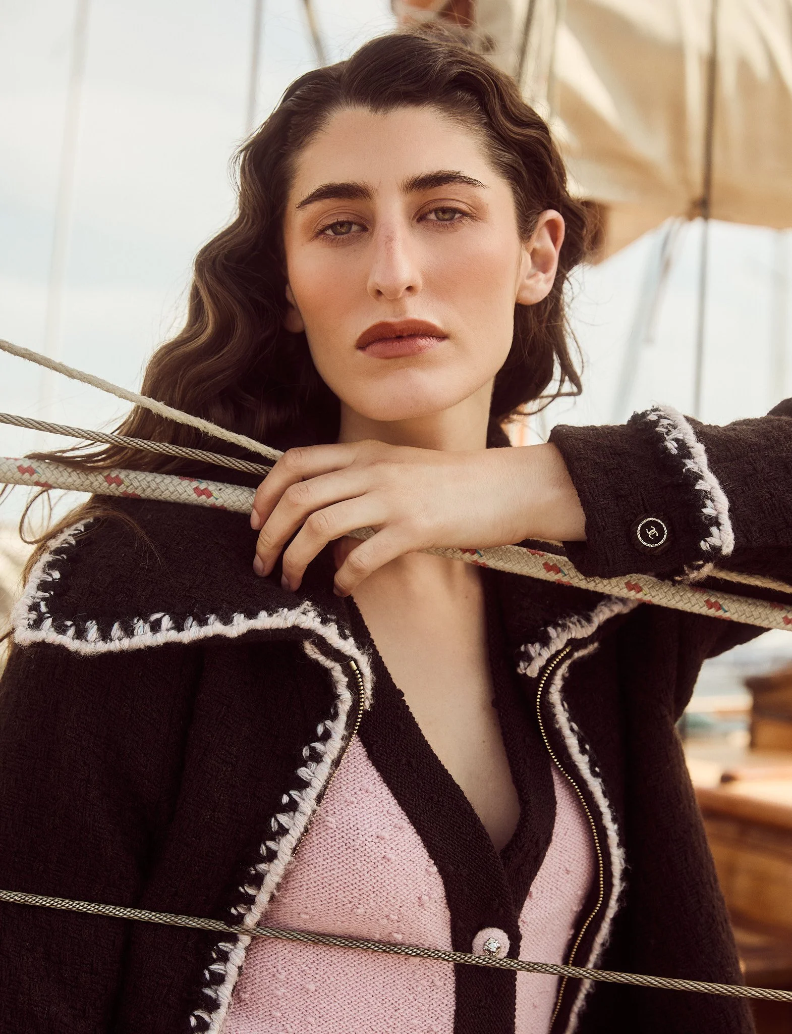 A woman with wavy brown hair and light makeup gazes at the camera, holding a rope draped across her shoulders in an outdoor setting with a sailboat and cream-colored sails in the background.