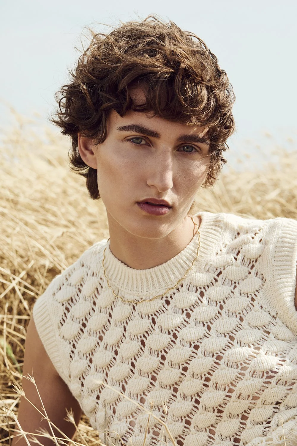 Young woman with curly brown hair and fair skin, wearing a cream-colored knitted sleeveless top with a gold chain necklace, standing in a field of dry grass under a clear sky.