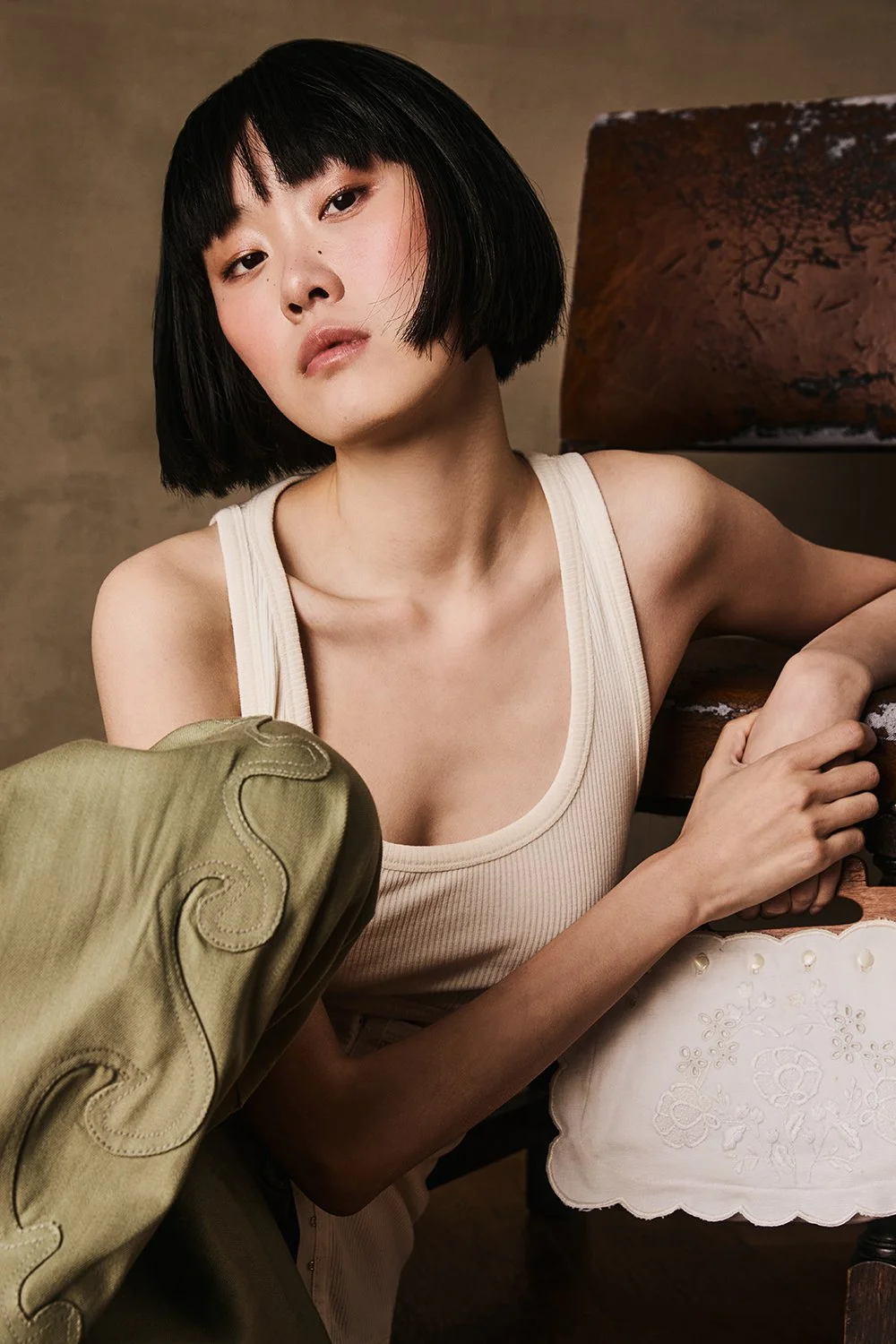 A woman with short black hair, wearing a beige sleeveless top, sitting next to a wooden table and a vintage lamp in a warmly lit setting.