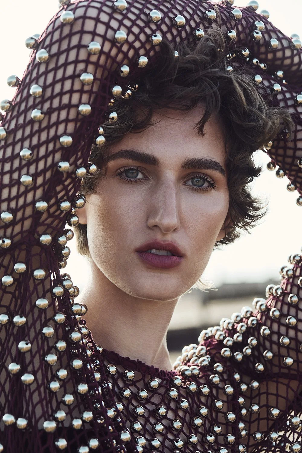 A young woman with short wavy hair and striking blue eyes, wearing a black netted dress adorned with silver beads, holding her arms above her head outdoors.