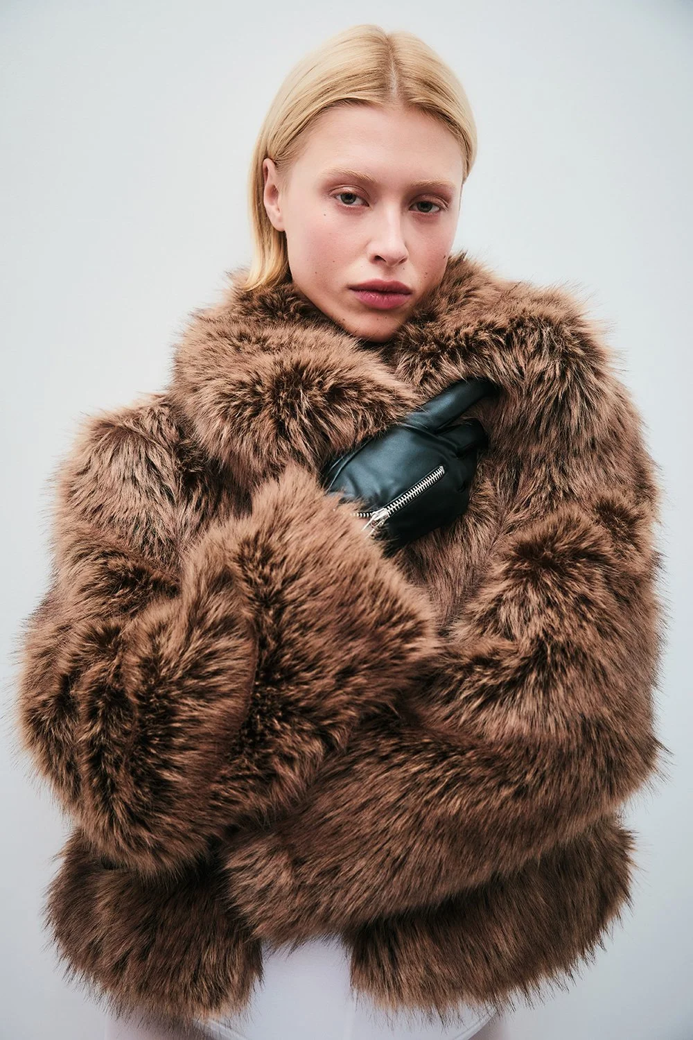 A woman with blonde hair wearing a large brown faux fur coat and black leather glove, holding her coat collar with her right hand, against a plain light-colored background.