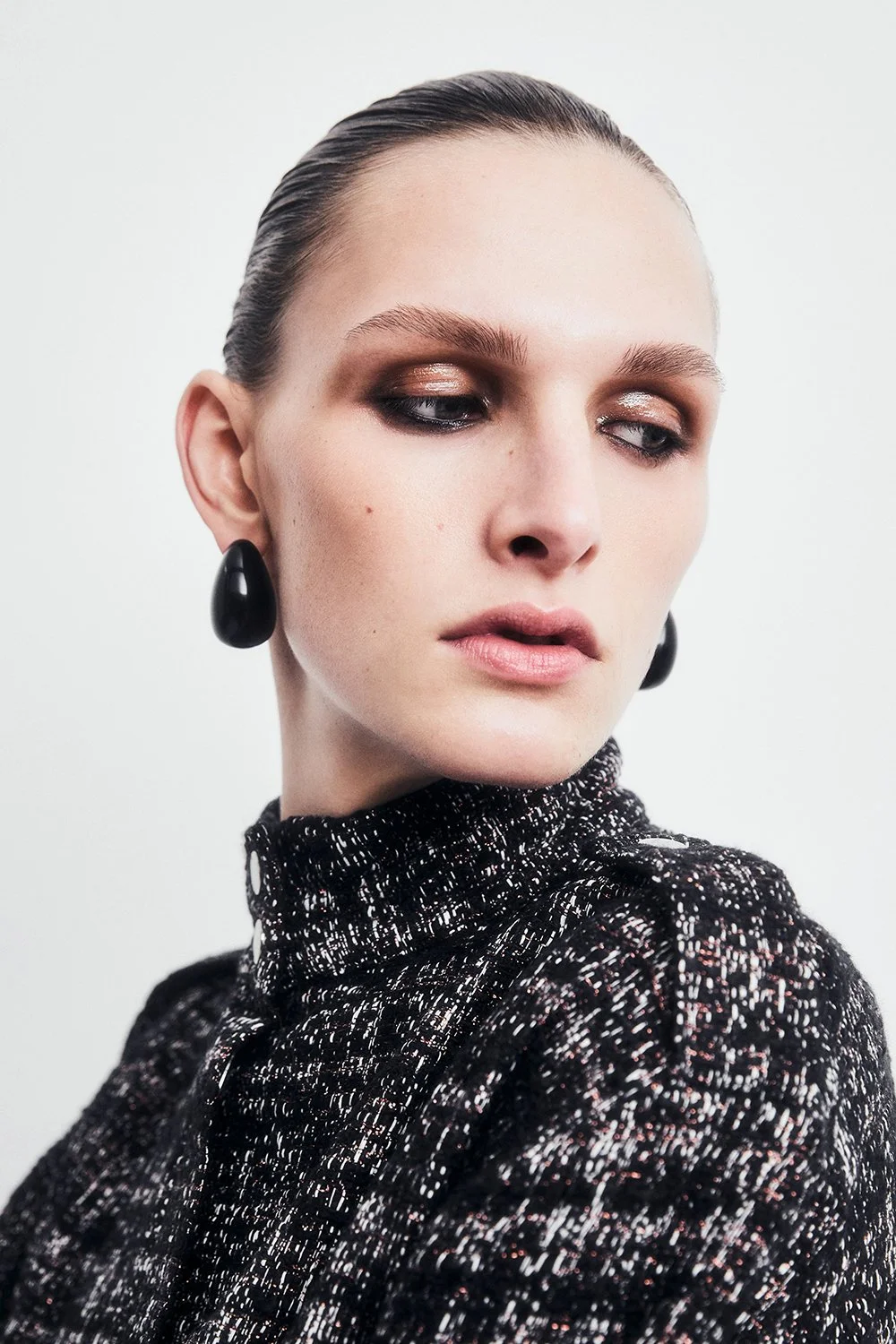 A woman with blonde hair slicked back, wearing black earrings and a black and white textured high-neck top.