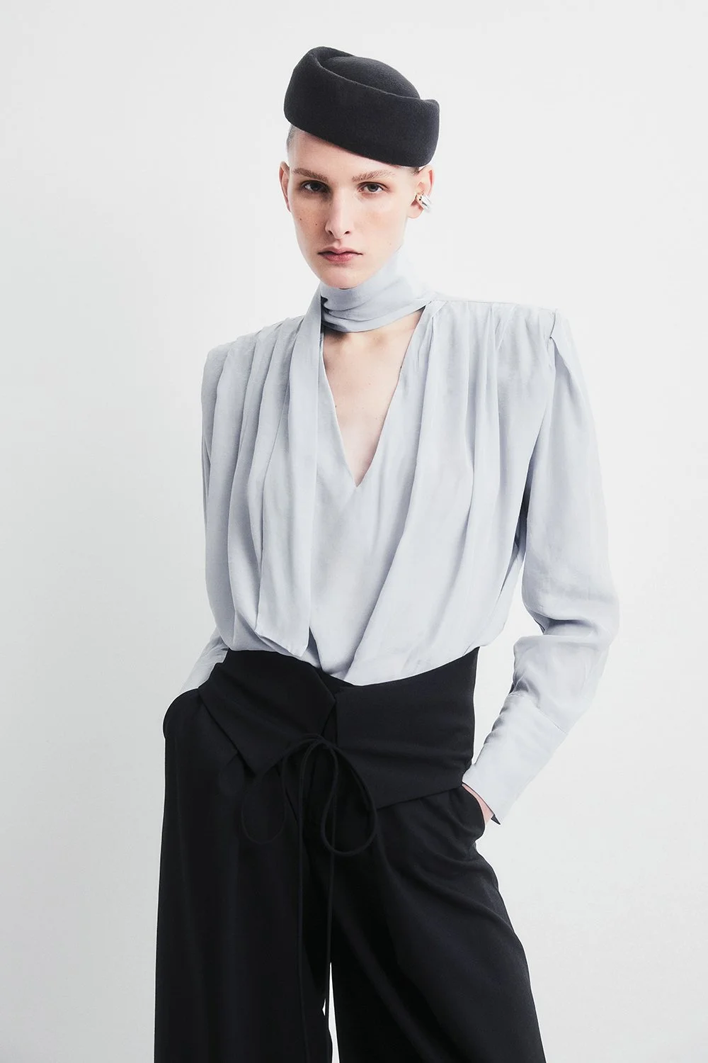 A woman with short blonde hair wearing a black hat, a light gray blouse, and black pants against a plain white background.