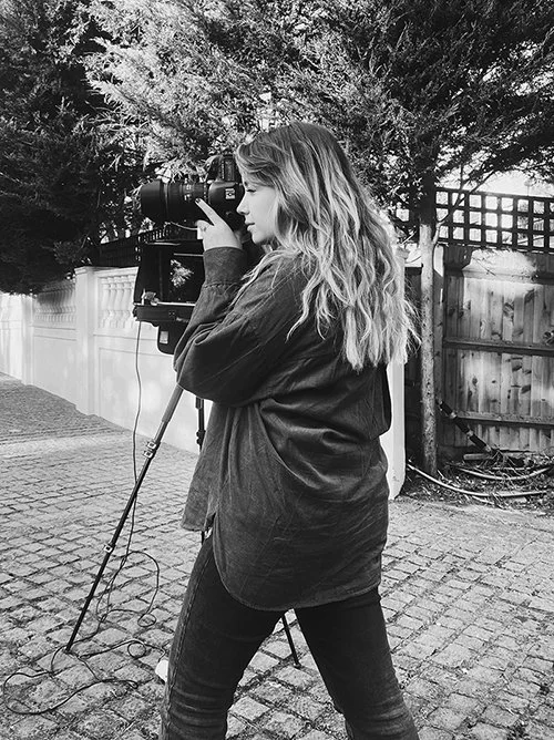 A woman with long hair takes a photo outdoors using a camera mounted on a tripod amid trees and a wooden fence.