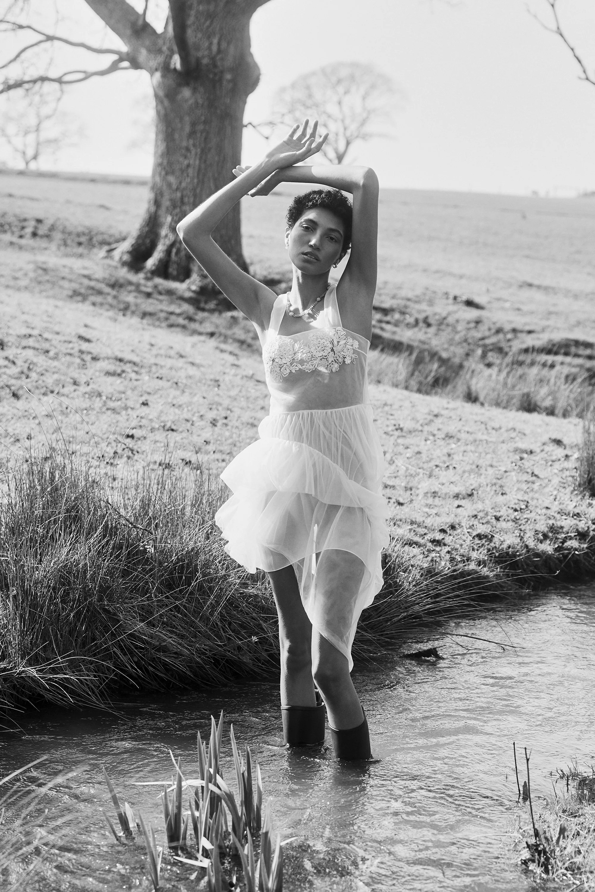 A woman in a light, lace dress standing in a creek with tall grass and trees in the background, striking a pose with arms raised above her head.