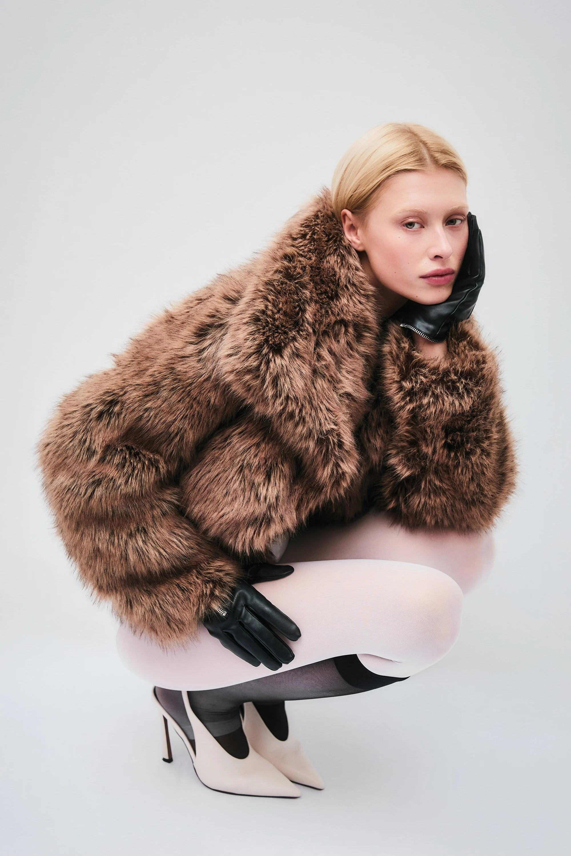A woman kneeling in a fashion pose, wearing a brown faux fur coat, black gloves, light pink tights, and high-heeled black and white shoes, against a plain white background.