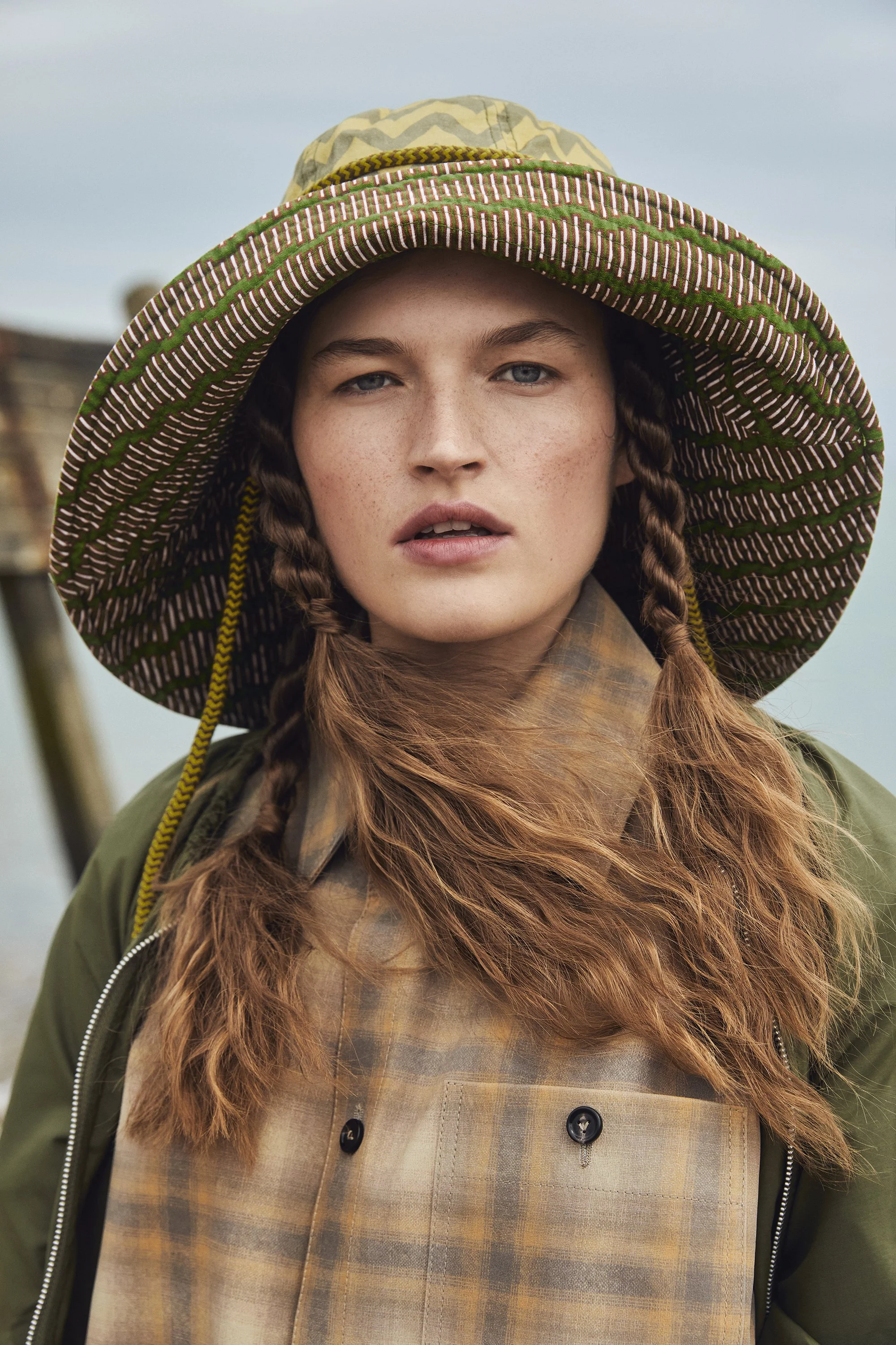 A young woman with braided hair wearing a large, wide-brimmed patterned hat and outdoor clothing, standing outdoors against a cloudy sky.
