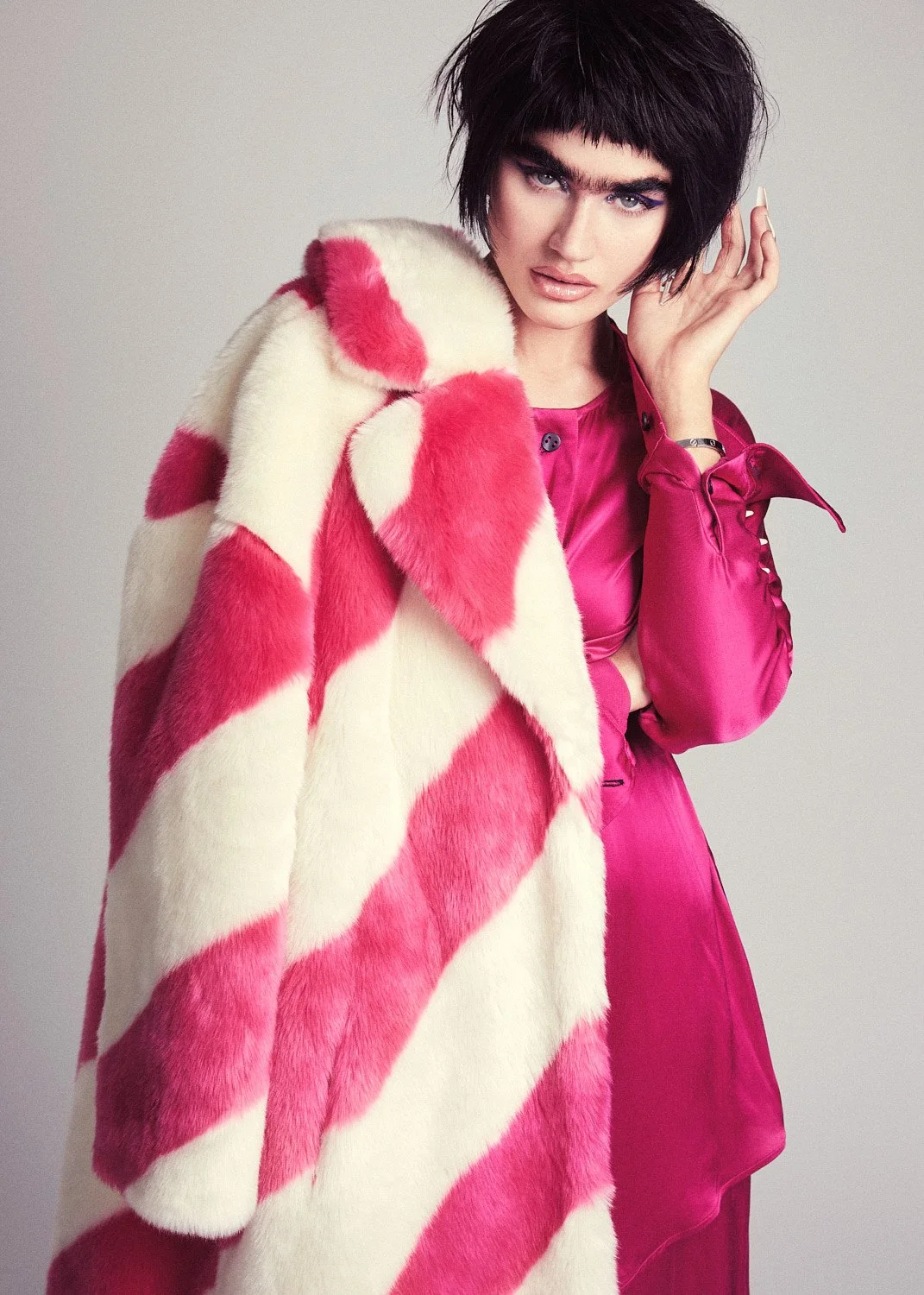 A woman with dark, short, messy hair and bold makeup wears a pink satin shirt and a pink and cream striped fur coat.