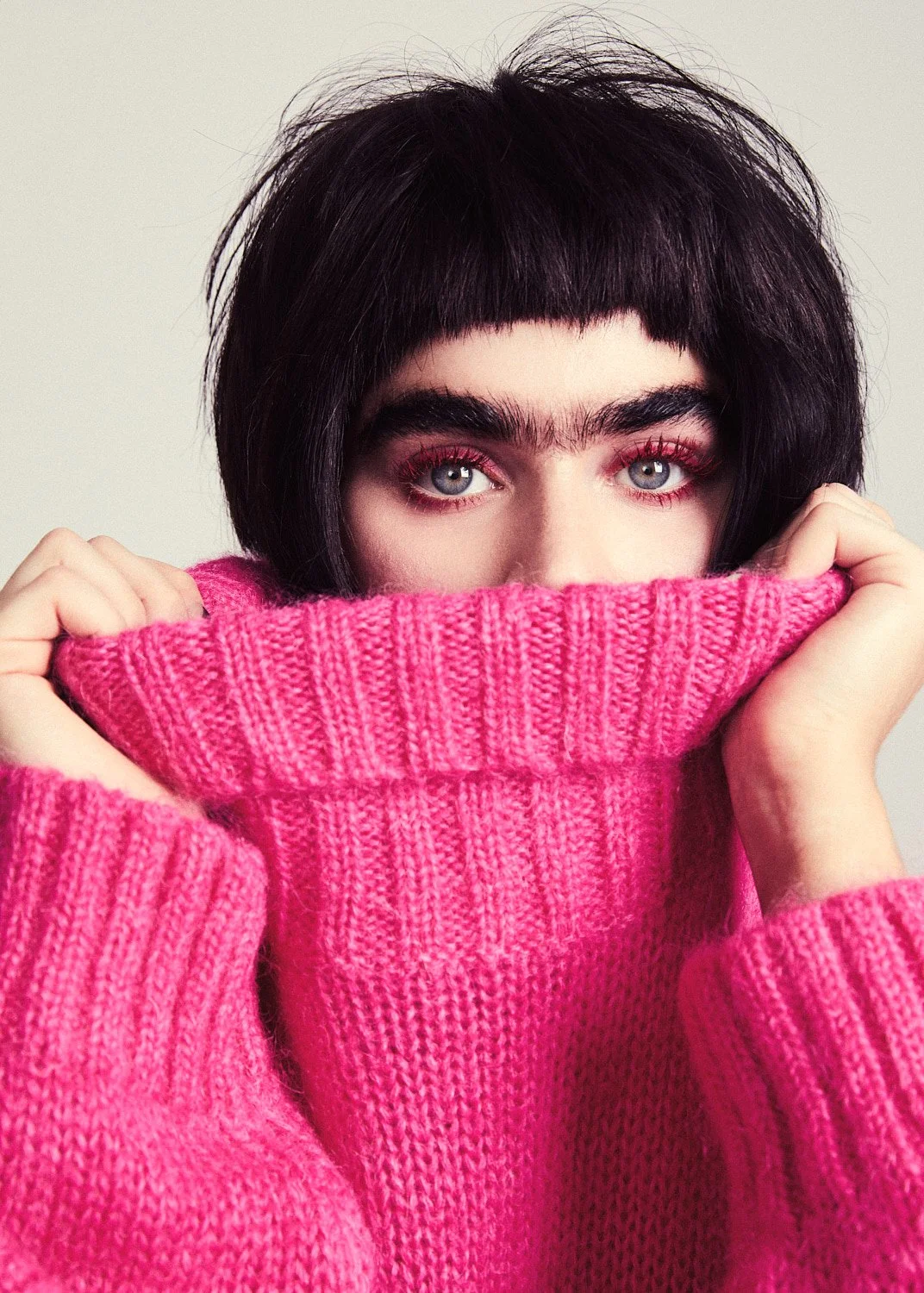 A person with dark short hair and bright blue eyes, wearing pink eyeshadow, holding a pink knitted sweater up to cover the lower part of their face.