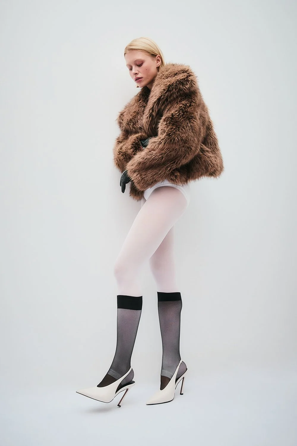 A woman wearing a brown fur coat, white tights, sheer black and gray stockings, and white high-heeled shoes stands against a plain light background.