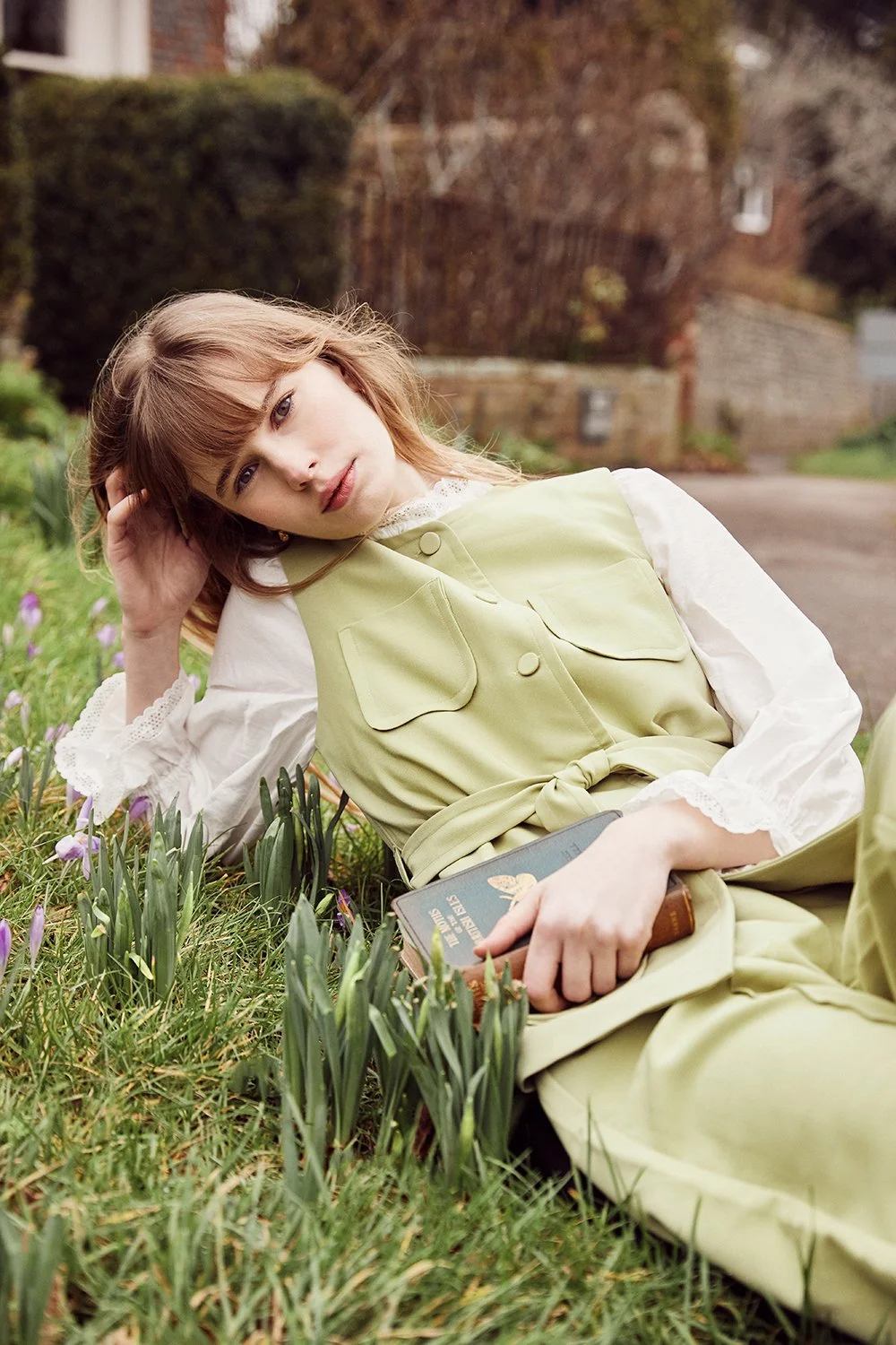 A young woman with reddish hair lying on the grass in a garden with purple flowers, holding a book and looking at the camera.