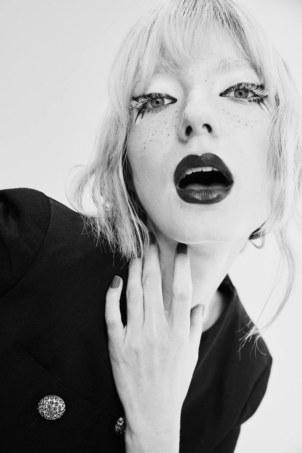 A black and white close-up photograph of a woman with short, wavy light-colored hair, dark lipstick, and dramatic eye makeup. She has an expressive facial expression with her mouth slightly open and her hand touching her neck, wearing a dark jacket w