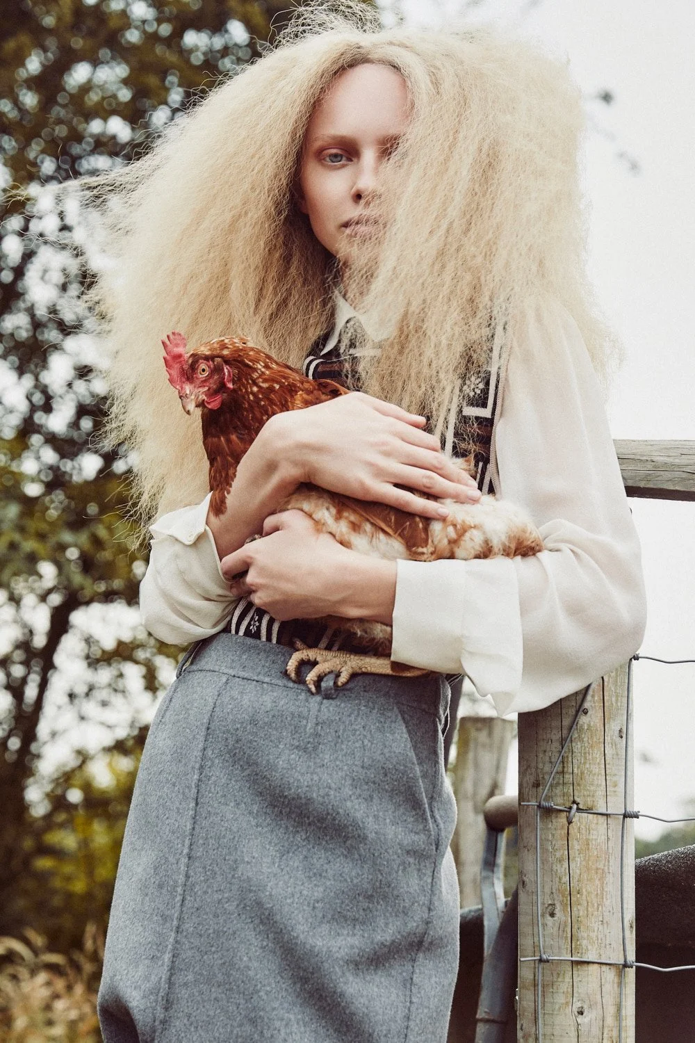 A woman with long, blonde, frizzy hair holding a hen outdoors, standing near a wooden fence post.