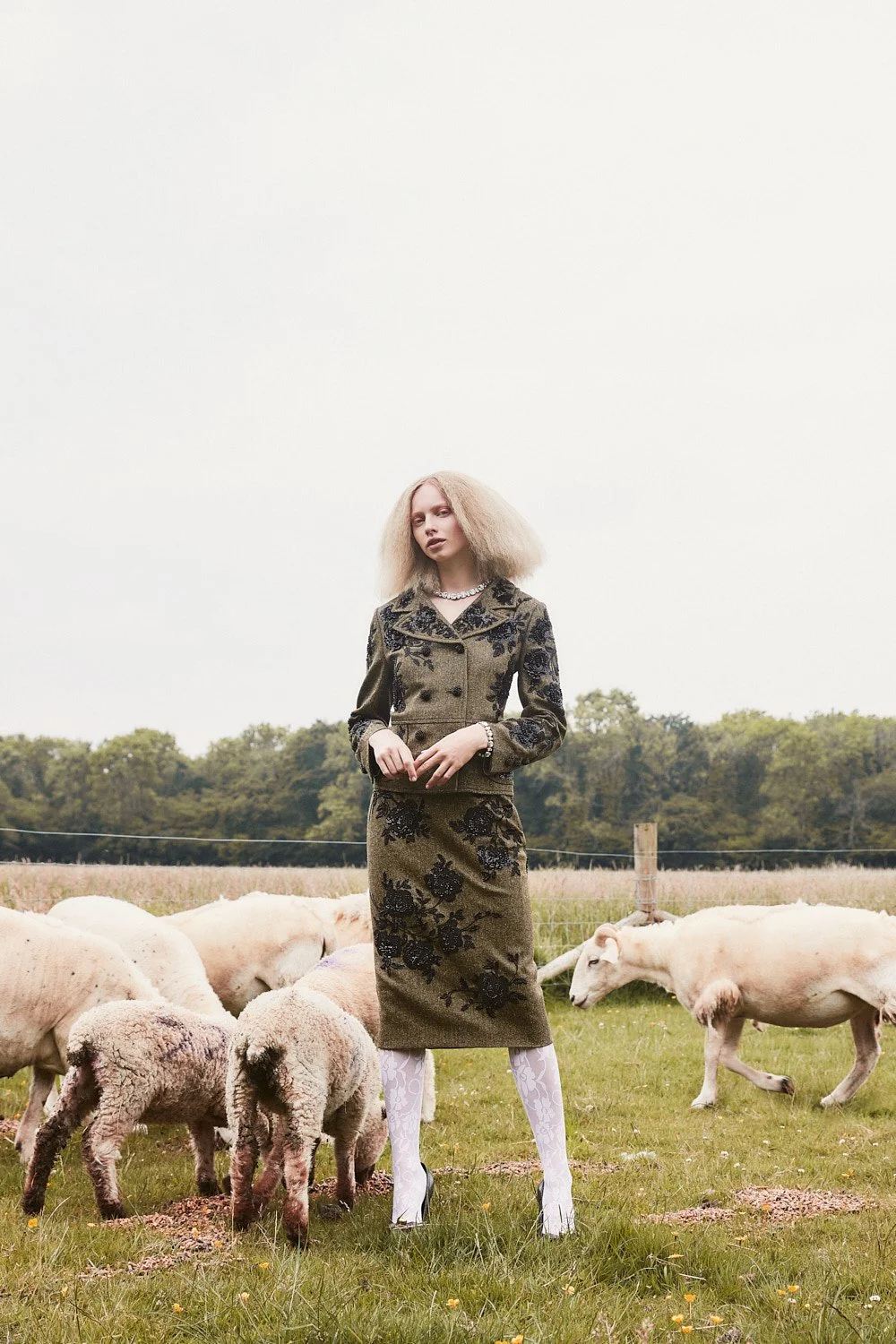 A woman dressed in a patterned skirt and jacket standing in a field surrounded by sheep and goats.