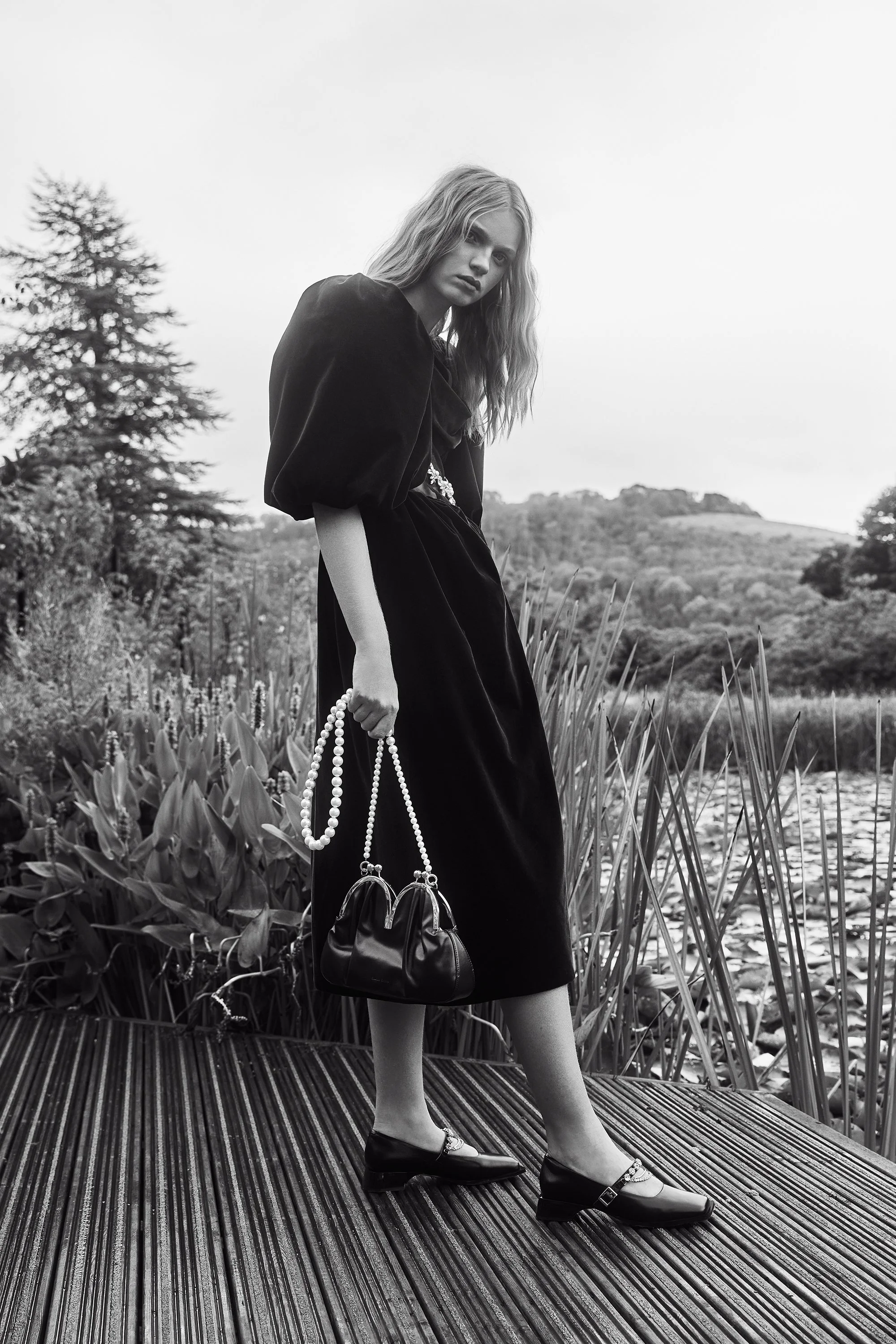 A woman in a black dress holding a handbag with a pearl strap, standing outdoors on a wooden platform with plants and hills in the background.