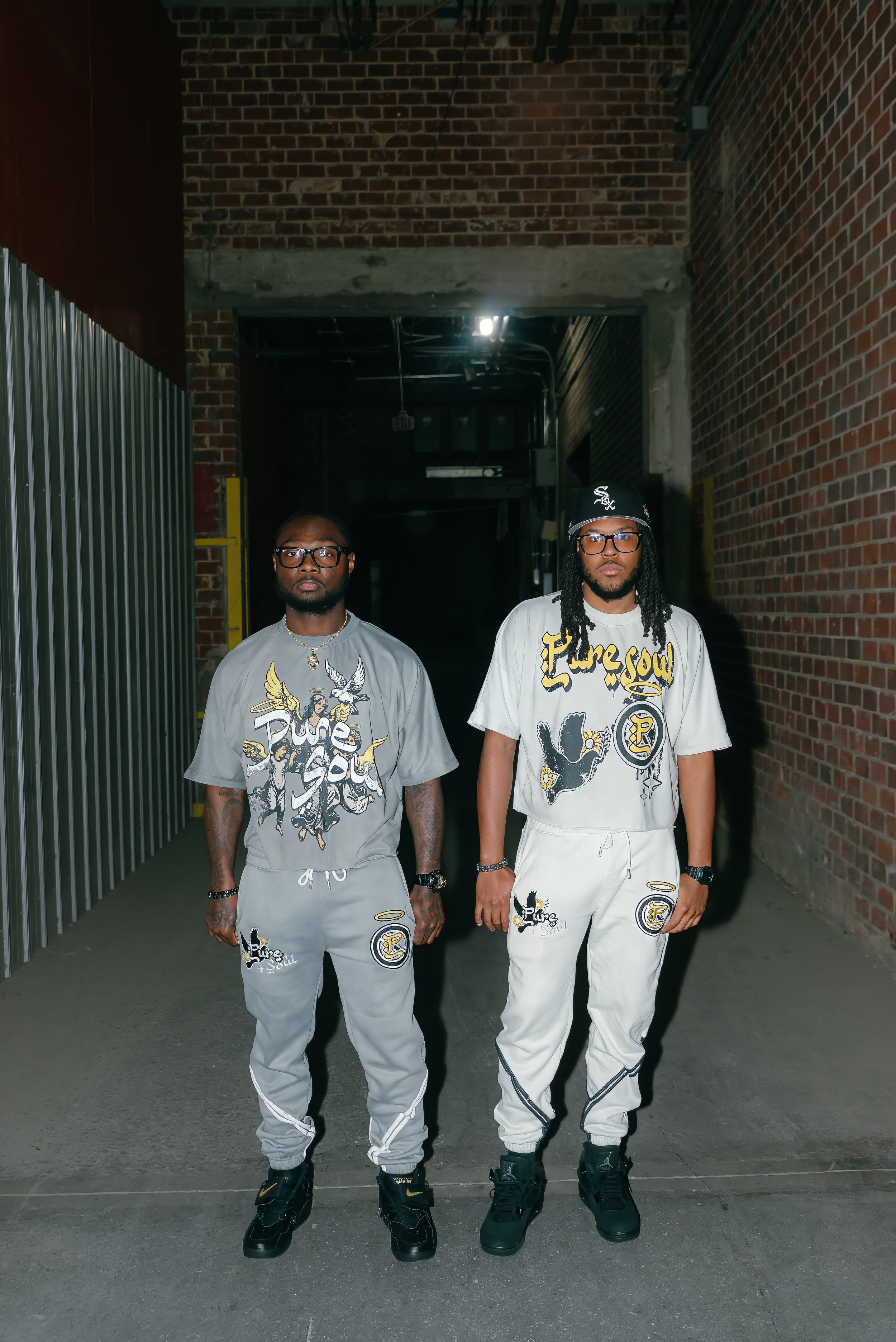 Two men standing side by side in an alley with brick walls, wearing matching streetwear outfits with graphic designs and sneakers.