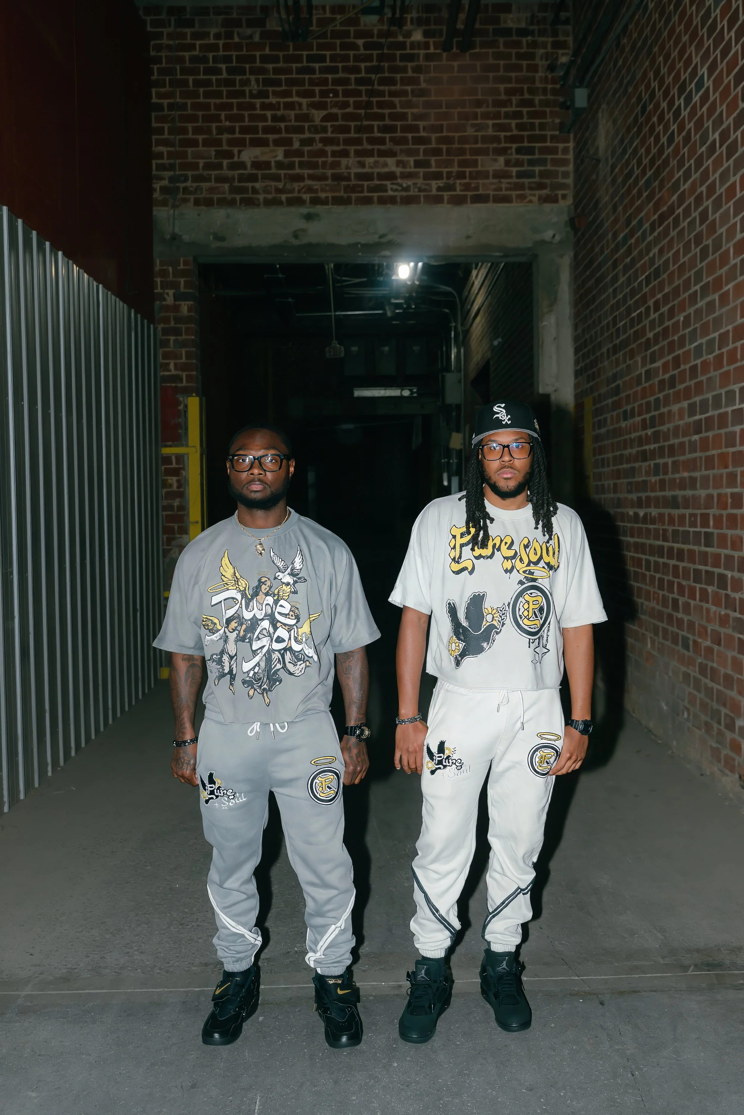 Two men standing in an alleyway wearing matching streetwear outfits with graphic designs, graffiti style text, and sneakers.