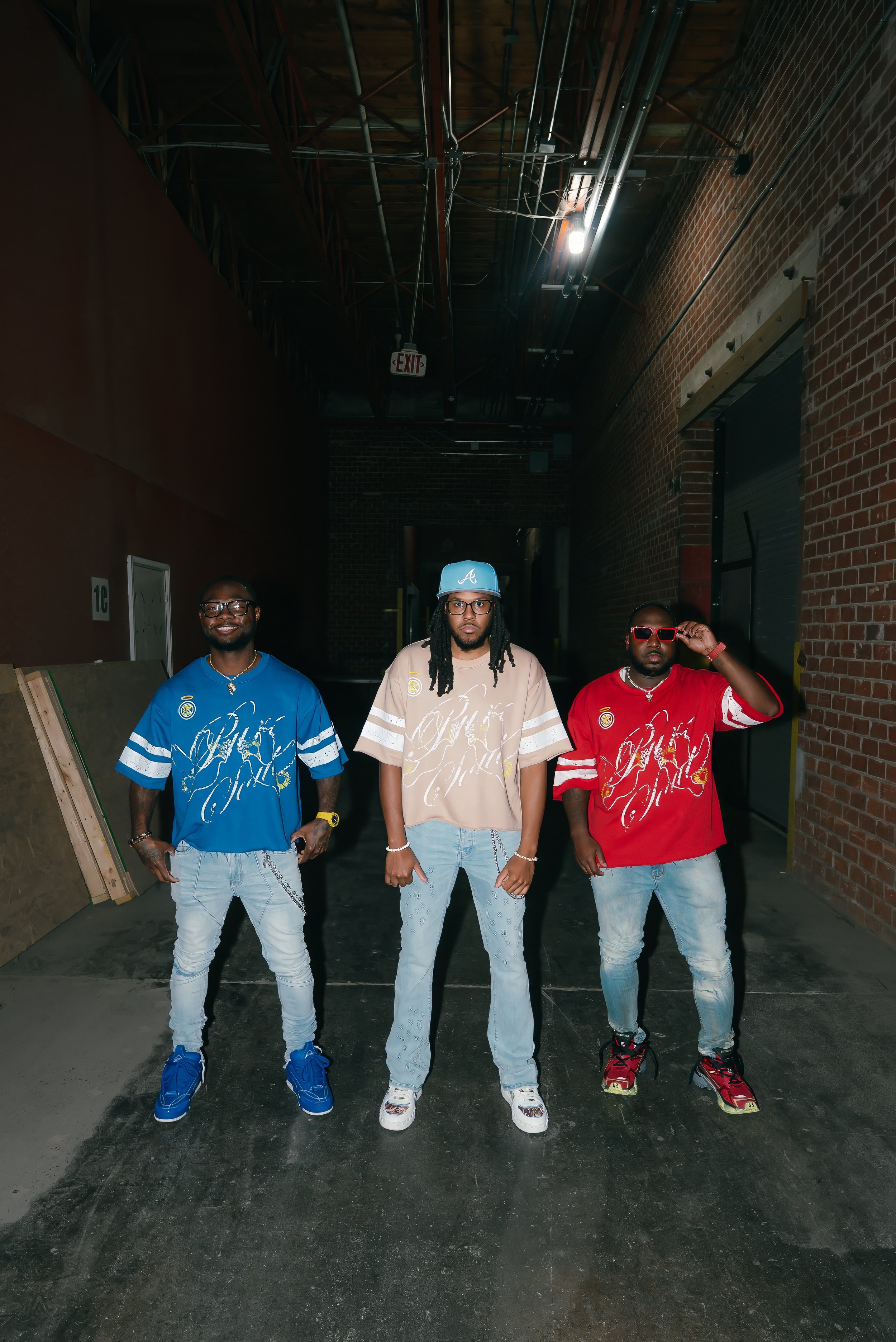 Three men standing in a dimly lit alleyway with brick walls, wearing colorful oversized shirts, jeans, and sneakers, posing confidently for the camera.