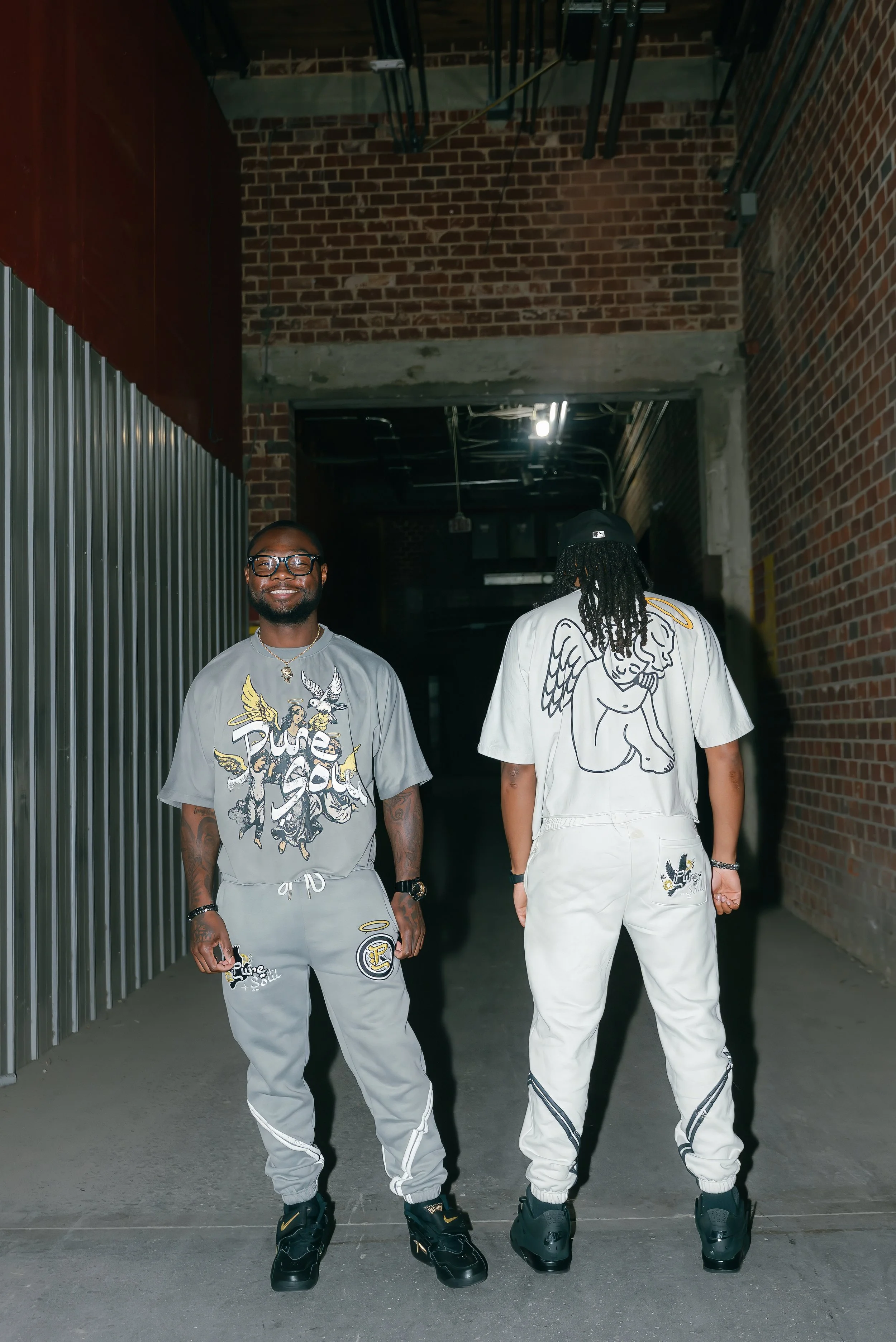Two men standing in a brick and metal corridor with graffiti on their clothing, one facing forward and smiling, the other with his back turned.